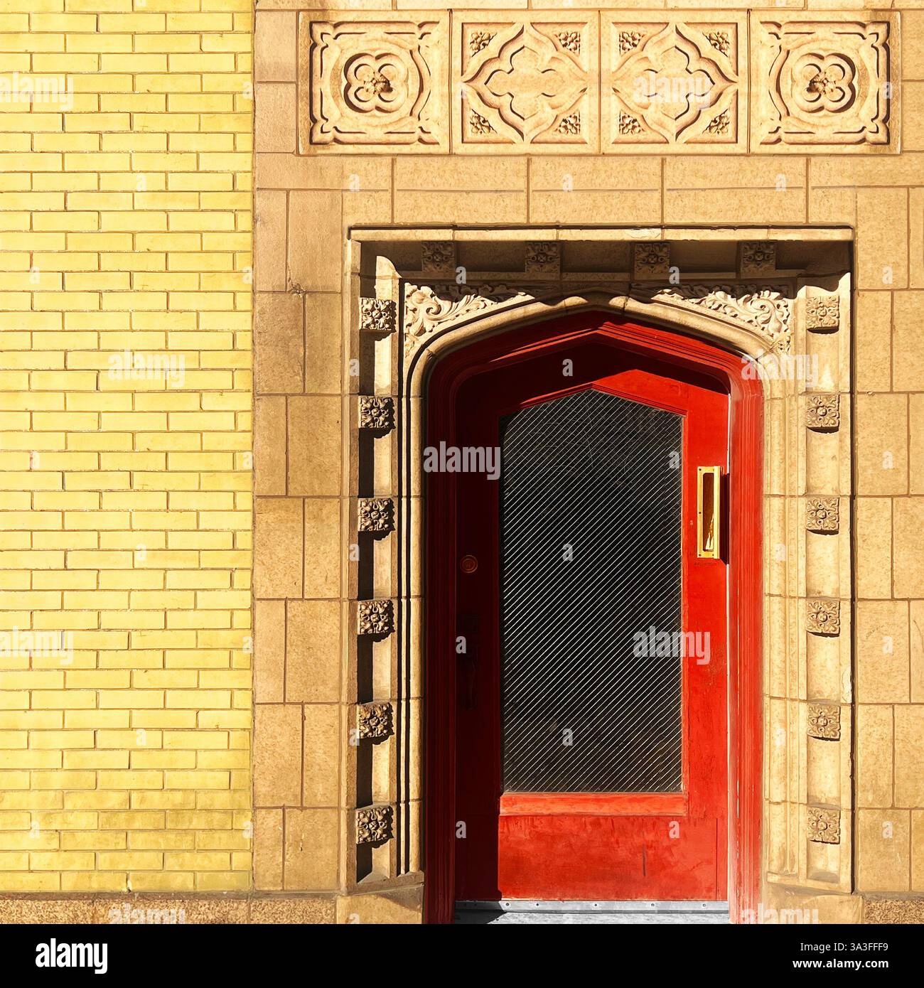 Porta ad arco rossa con un pannello di vetro testurizzato, incorniciato da intricate incisioni in pietra su un edificio in mattoni gialli a Chicago - Immagine stock catturata con smartphone