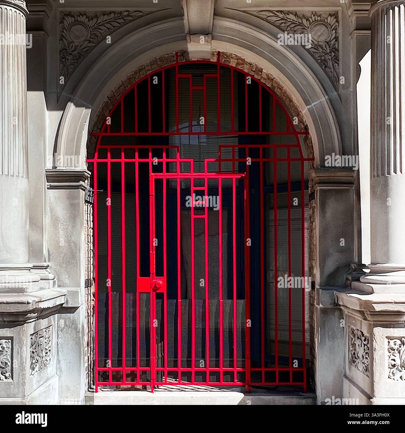 Cancello in ferro battuto rosso dal design geometrico, situato all'interno di un grande ingresso in pietra arcuata fiancheggiato da colonne classiche e intricate incisioni a Chicago - Immagine stock catturata con smartphone