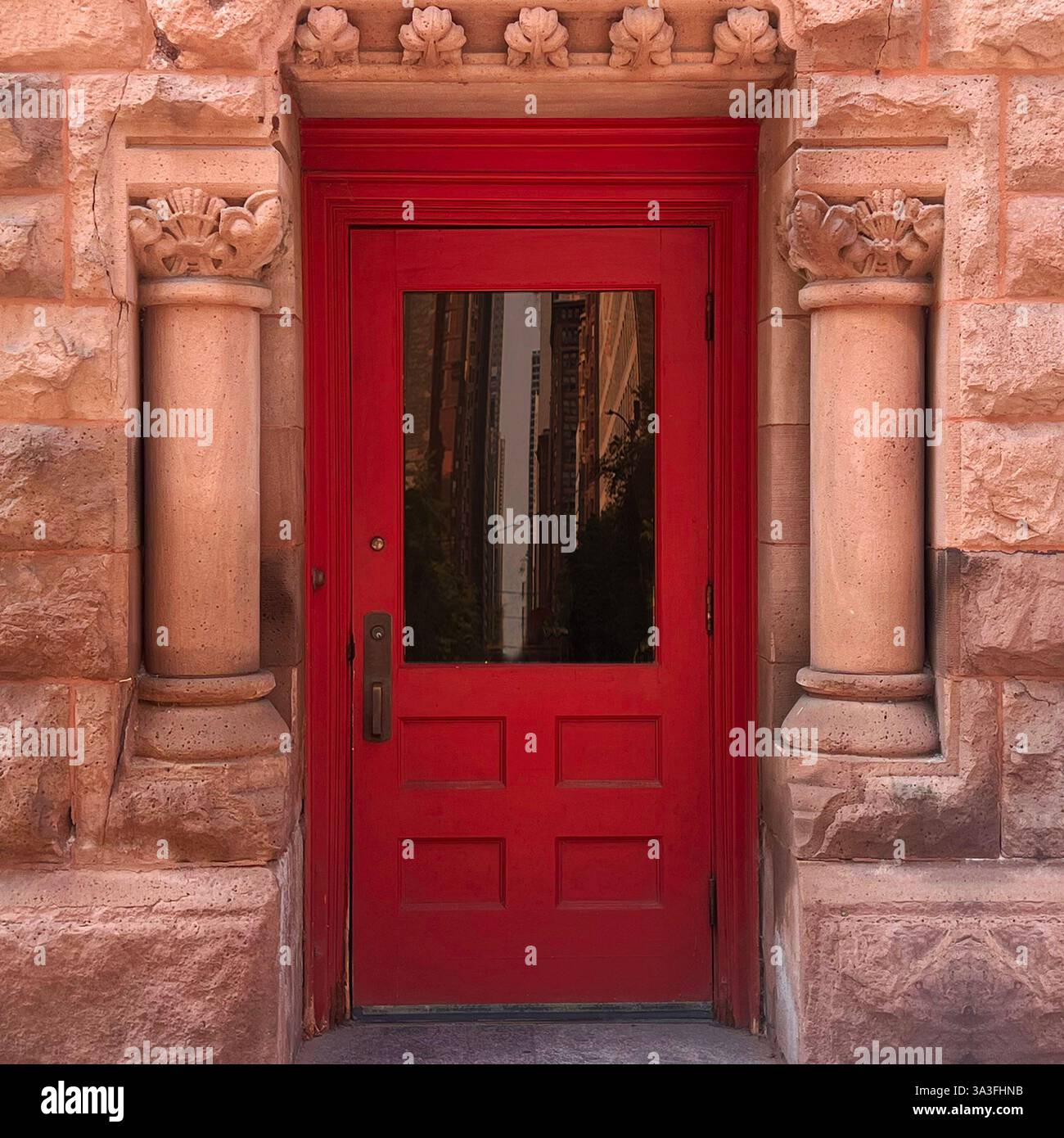 Porta di legno rossa con un pannello di vetro riflettente, incorniciato da colonne di pietra rosa ornate e intricate incisioni, adagiata all'interno di un edificio storico urbano. - Immagine stock catturata con smartphone