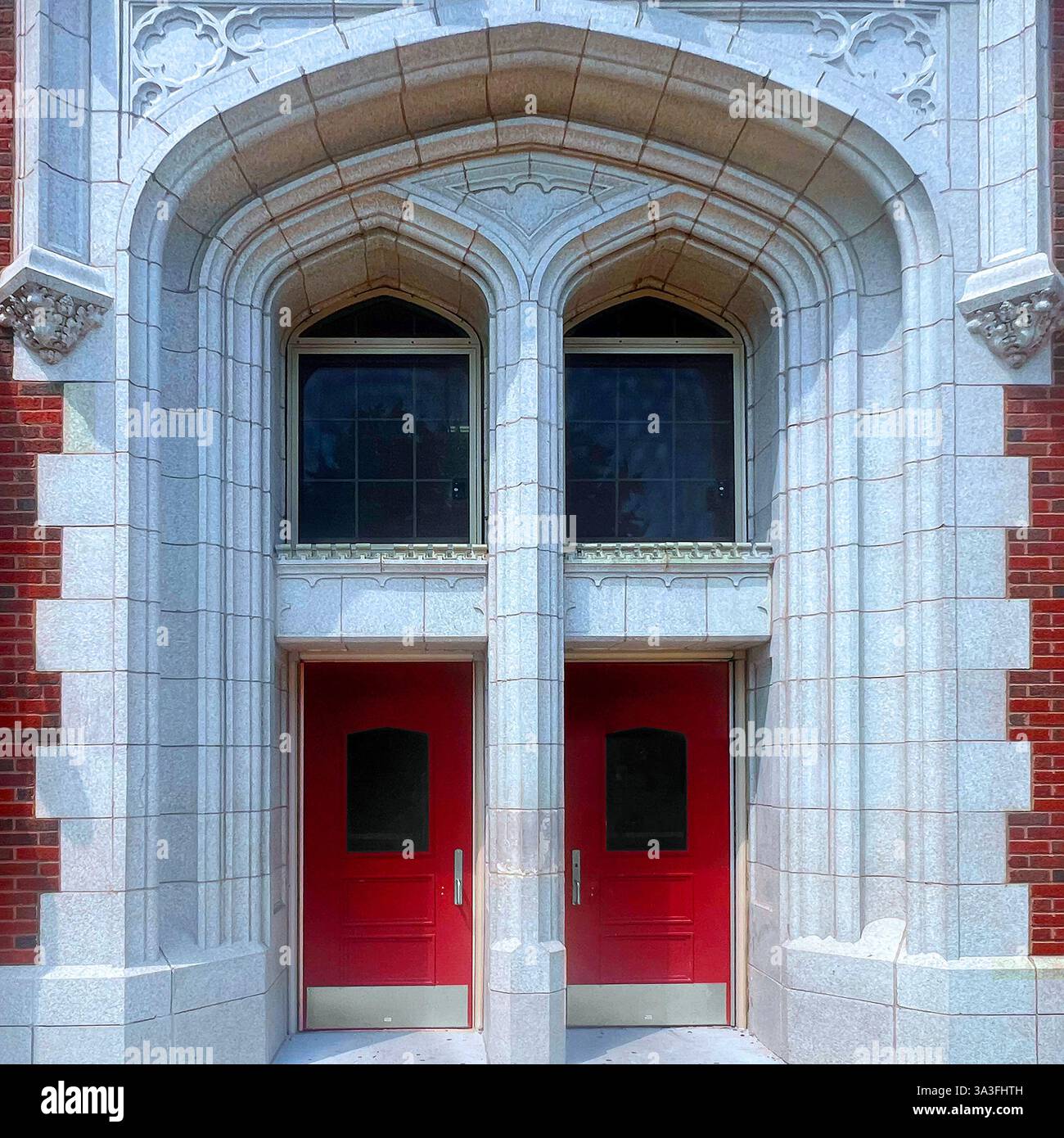 Un paio di porte rosse adagiate all'interno di un grande arco in pietra in stile gotico, incorniciato da intricati dettagli architettonici su uno storico edificio in mattoni. - Immagine stock catturata con smartphone