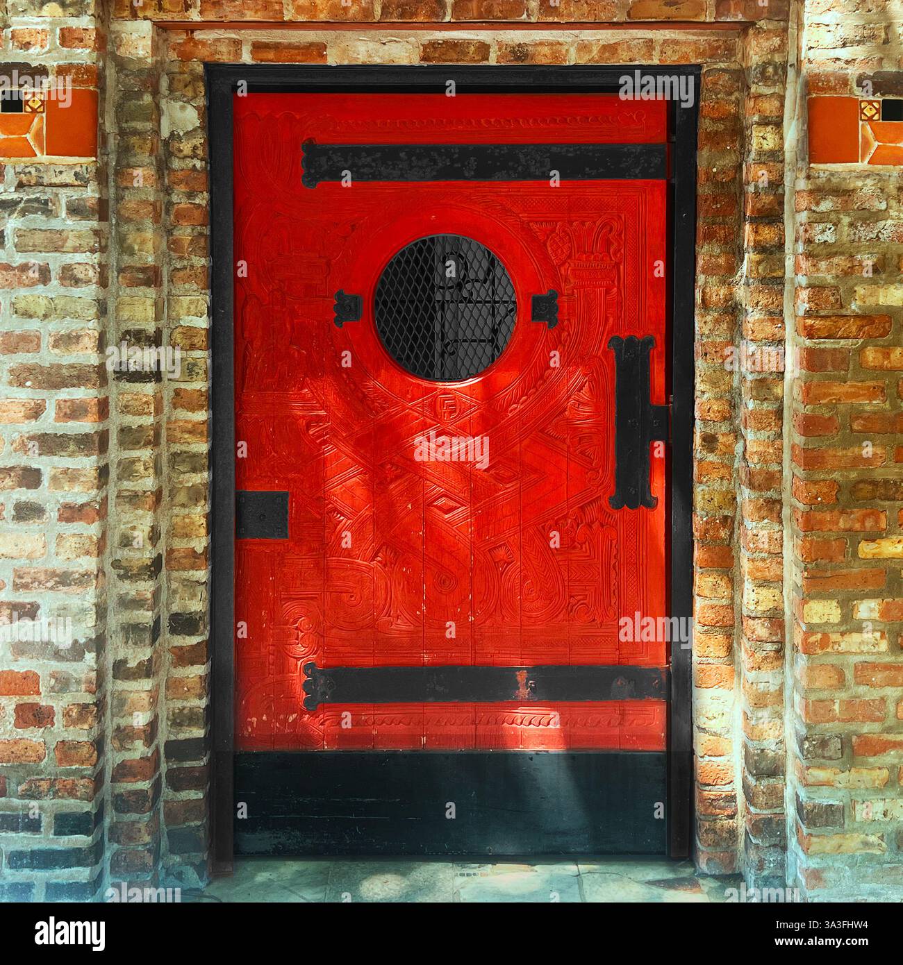 Porta in legno rosso Art Deco con intricate incisioni, ferramenta in ferro nero e una finestra circolare, collocata all'interno di una facciata in mattoni rustici a Chicago - Immagine stock catturata con smartphone