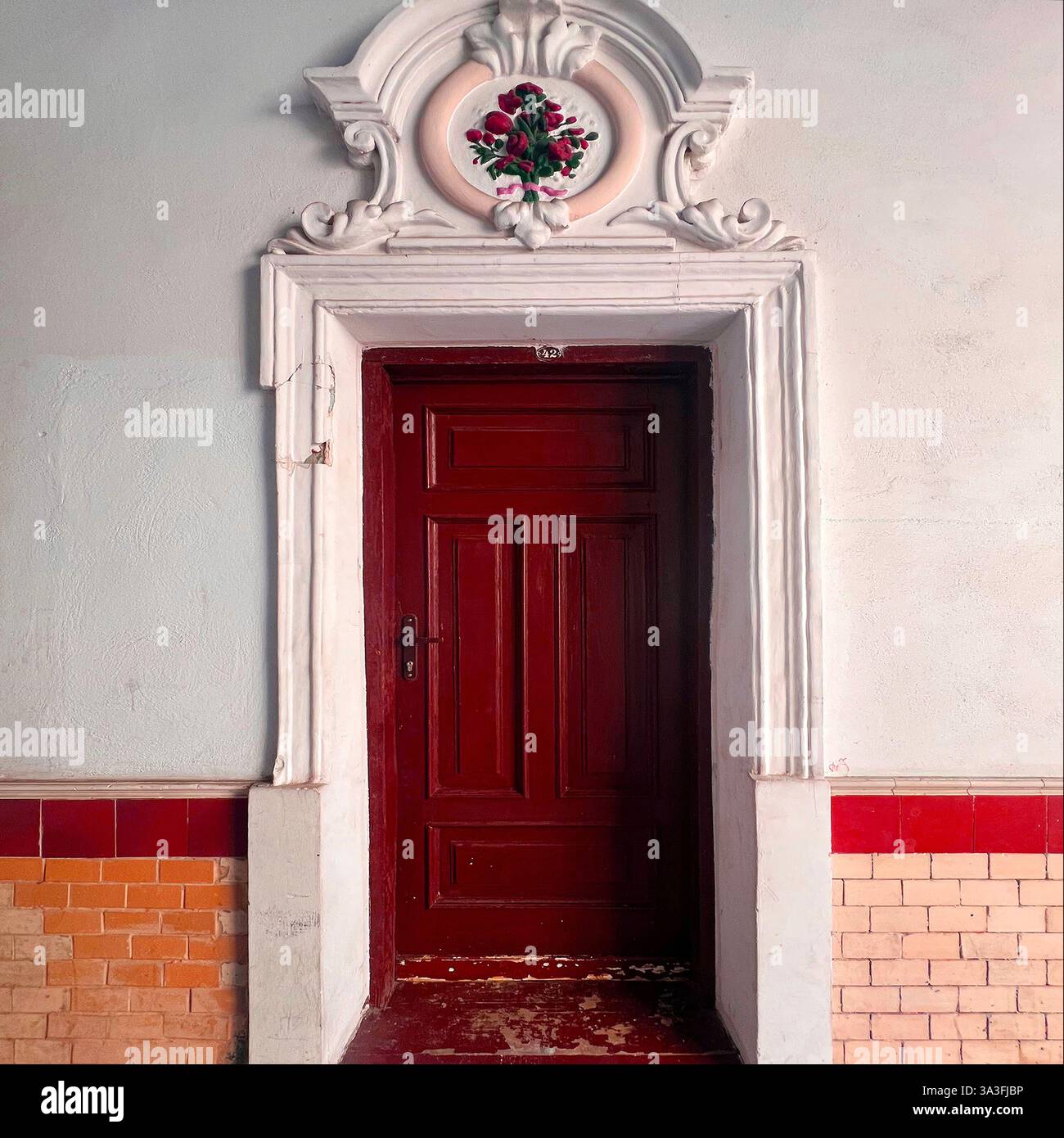 Una porta rossa ornata incorniciata da modanatura bianca decorativa e un rilievo floreale, situata in un edificio storico a Varsavia, Polonia - Immagine stock catturata con smartphone