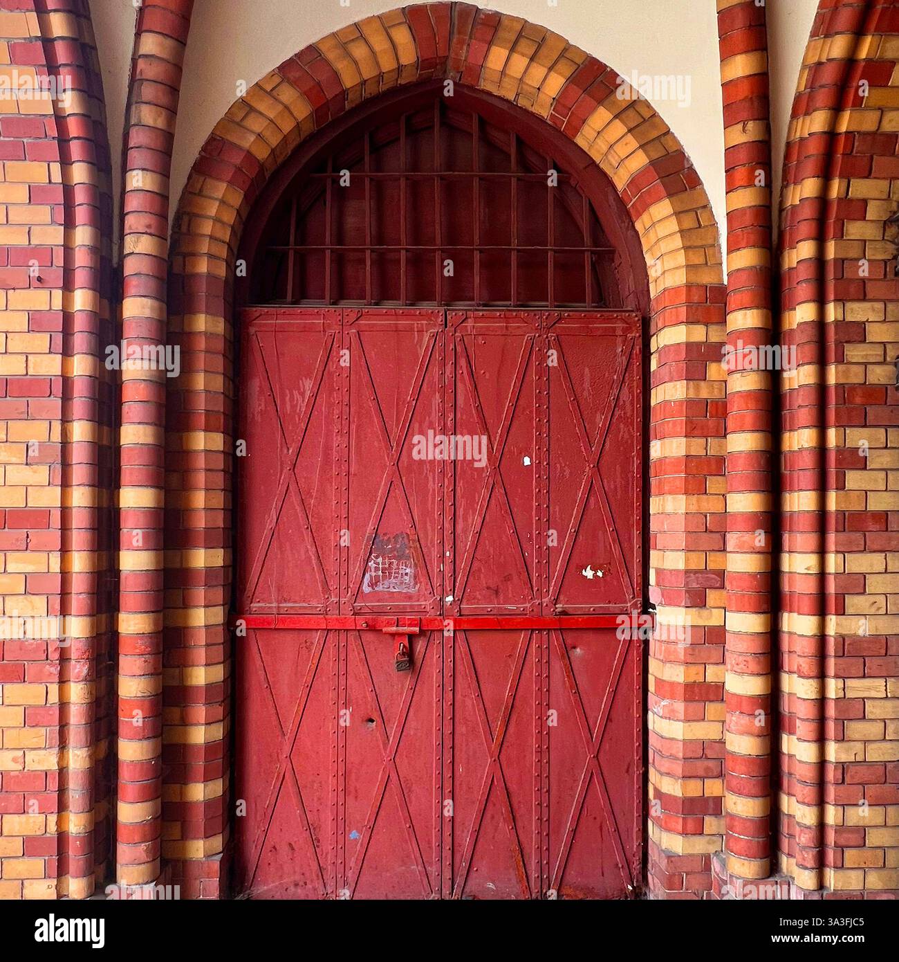 Una storica porta rossa in metallo con intricati motivi a diamante, adagiata all'interno di una facciata in mattoni in stile gotico a Varsavia, Polonia - Immagine stock catturata con smartphone