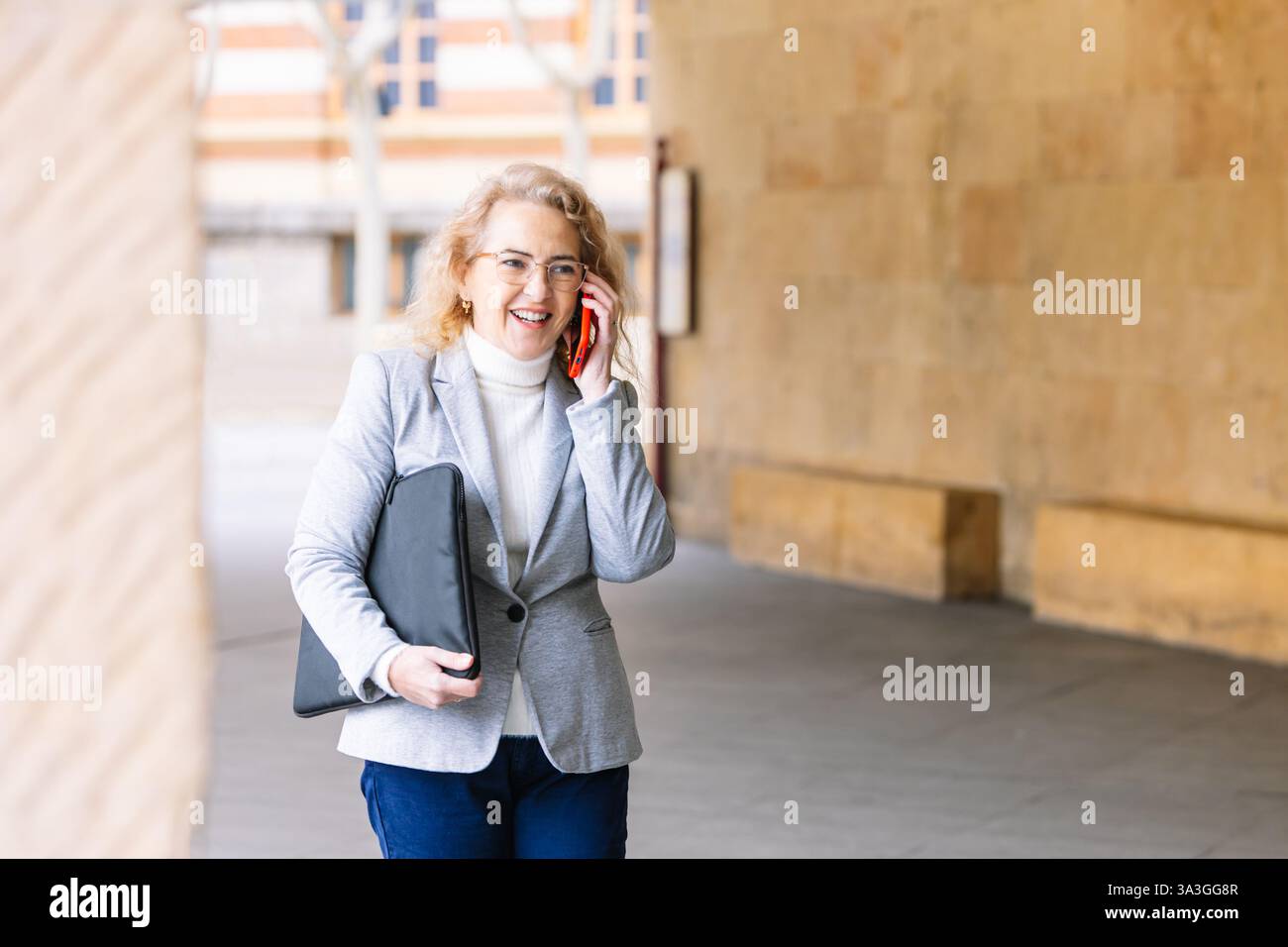 Una donna professionale con un blazer grigio e dolcevita bianca attraversa un ambiente urbano mentre parla con il suo smartphone. ha un portatile nero Foto Stock