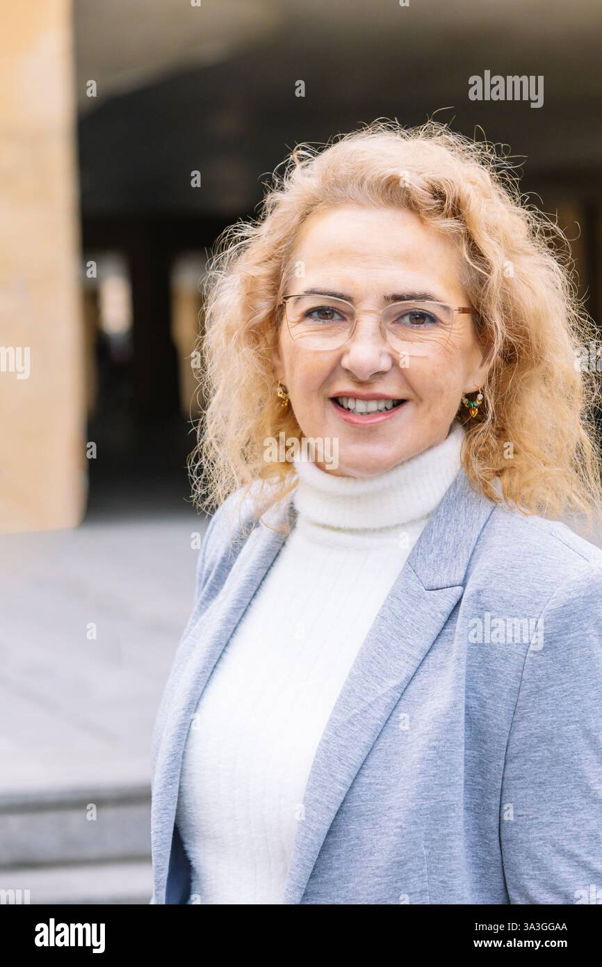 Donna matura professionale che indossa occhiali e un blazer grigio sopra un dolcevita bianco, sorridendo con sicurezza in un ambiente urbano. ideale per aziende e. Foto Stock