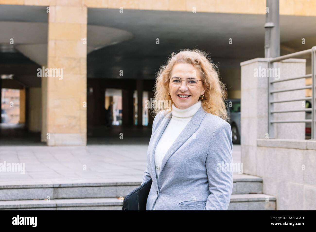 Donna professionale con occhiali e blazer grigio sorridente, mentre tiene in mano un portafoglio nero in un ambiente urbano. il suo atteggiamento fiducioso riflette il successo Foto Stock