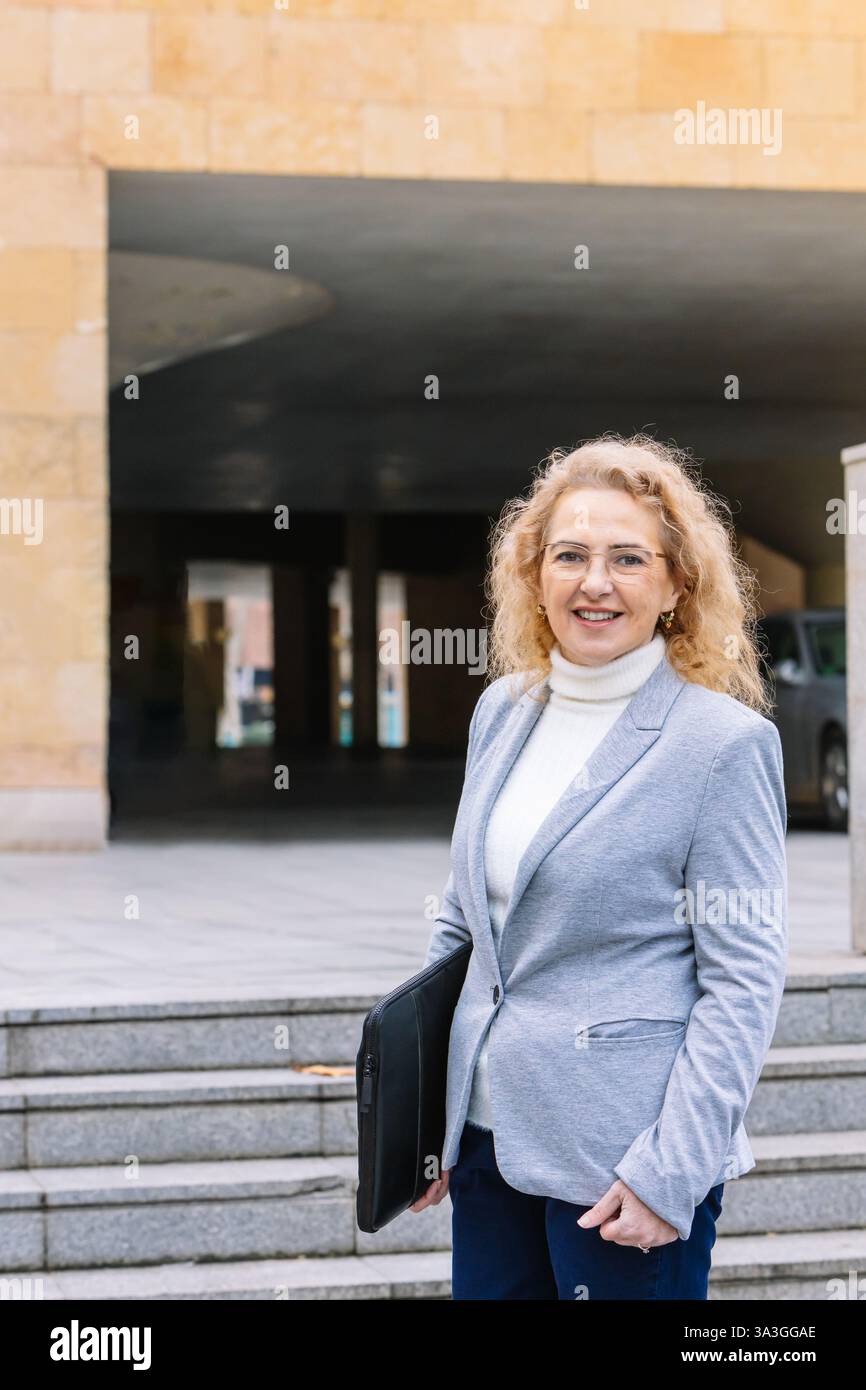 Donna professionista che indossa un blazer e occhiali grigi, sorridendo con sicurezza mentre ha in mano un portafoglio nero. si trova su una scala urbana, pronta per Foto Stock