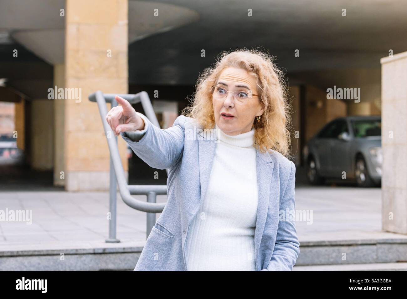 Donna d'affari matura ed espressiva con capelli e occhiali biondi ricci, con un blazer grigio chiaro e dolcevita bianco, che indica qualcosa con un sussulto Foto Stock