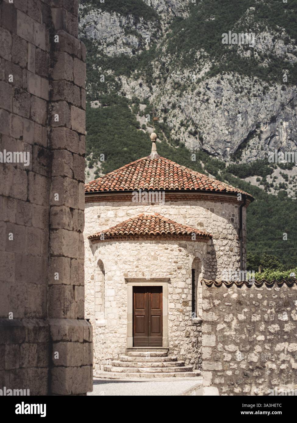 Una storica chiesa cripta in pietra a Venzone, Italia, con un'abside rotonda e tetto in terracotta, adagiata su uno sfondo di montagna spettacolare. Il sito è noto per Foto Stock