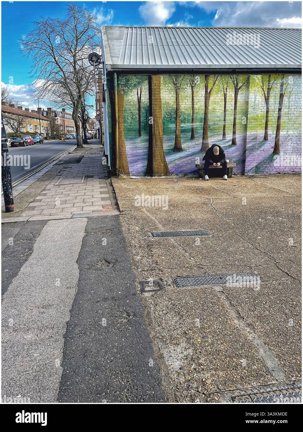 Giovane con un berretto da baseball, seduto su una panchina, a testa in giù guardando il suo cellulare. Sul muro dietro di lui c'è un grande murale di una foresta. Foto Stock