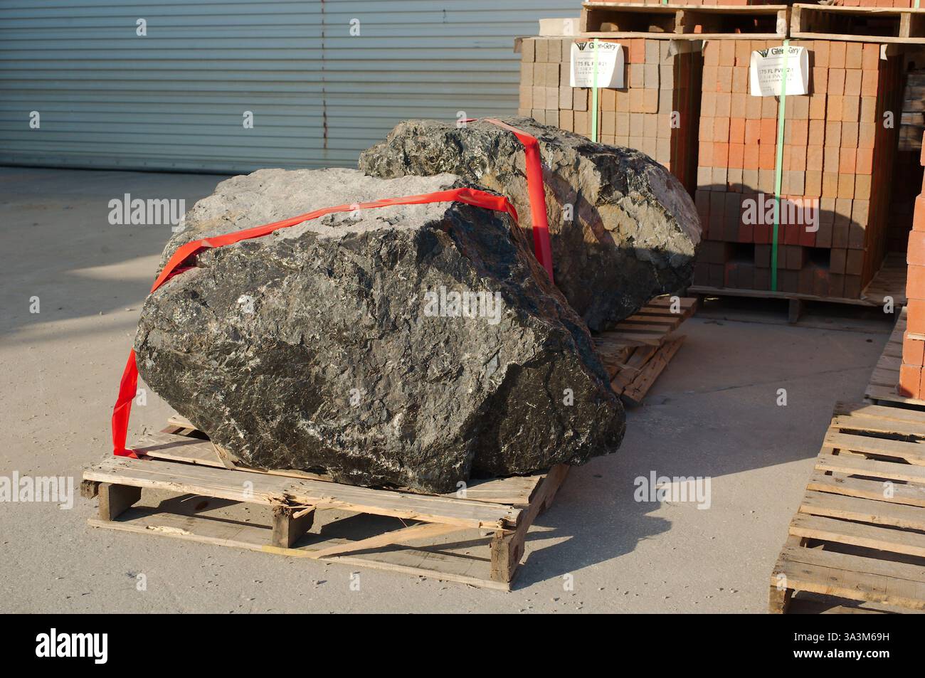 Grande masso di granito nero su una slitta di legno avvolta da cinghie arancioni. Sole nel tardo pomeriggio. Pallet di mattoni rossi nella parte posteriore su una lastra di cemento. Foto Stock