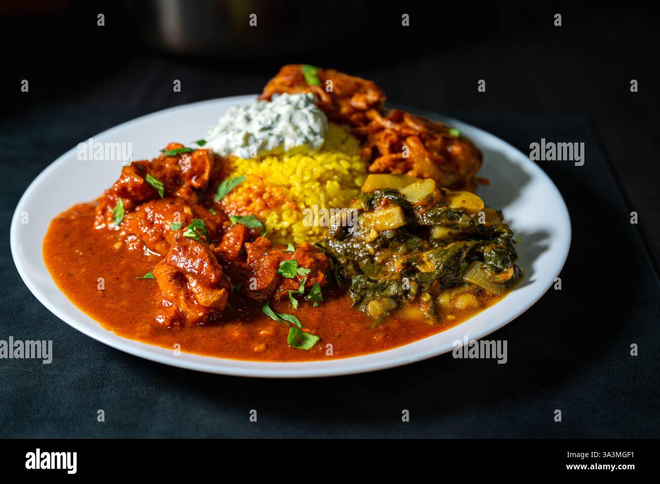 Pollo al burro indiano, noto anche come Murgh Makhani. Piatto colorato al curry servito con riso giallo, saag aloo, bhaji di cipolla e raita di cetrioli. Ben illuminato Foto Stock
