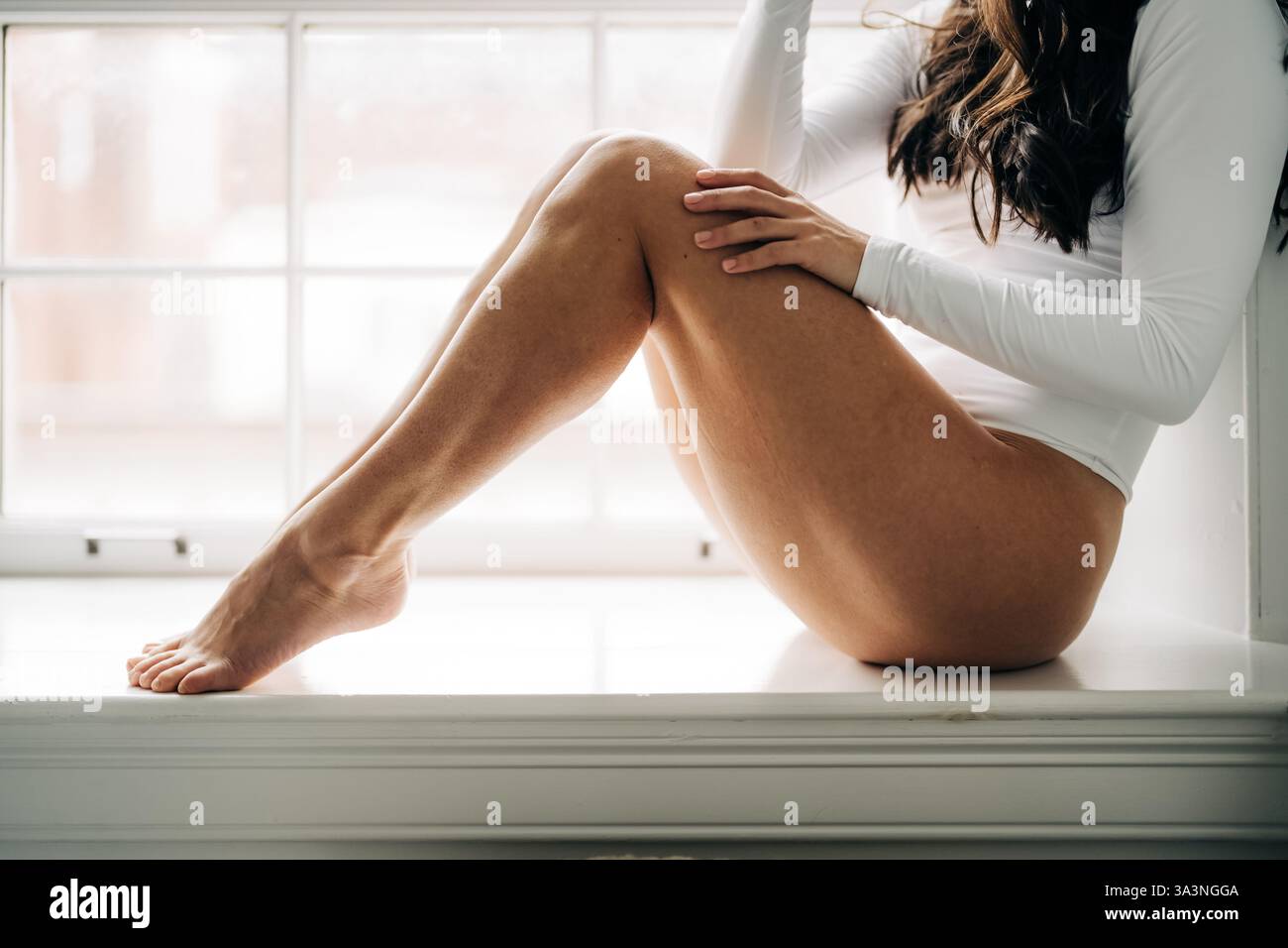 Primo piano delle gambe di una donna sedute da una finestra illuminata dal sole in bianco Foto Stock