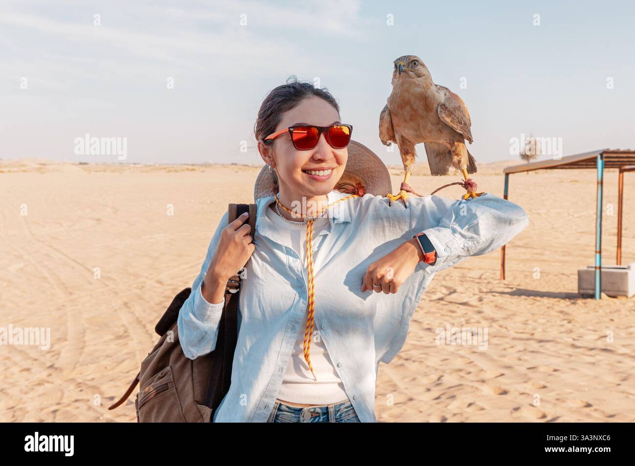 Giovane turista che indossa occhiali da sole, tiene un falco sul braccio, sorride nel deserto Foto Stock