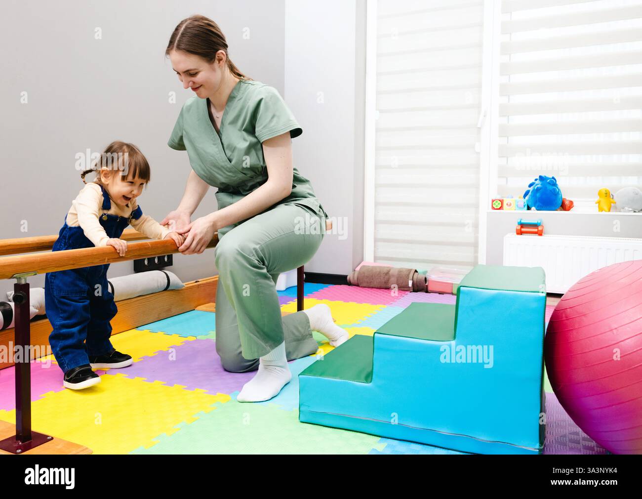 Giovane ragazza carina con paralisi cerebrale pratica in piedi e in equilibrio utilizzando corrimano con l'assistenza di fisioterapista in un colorato roo terapeutico Foto Stock