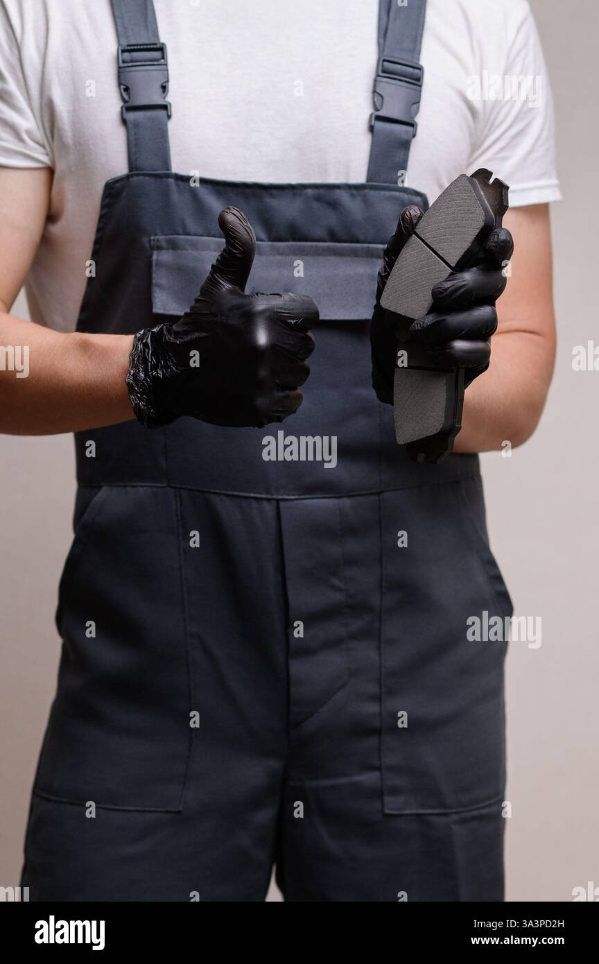 Il meccanico automatico in tuta tiene in mano due pastiglie dei freni, mostra il pollice in alto, primo piano. Concetto di sostituzione dei ricambi auto, manutenzione della vettura, facilità di manutenzione dell'impianto frenante. Verticale Foto Stock