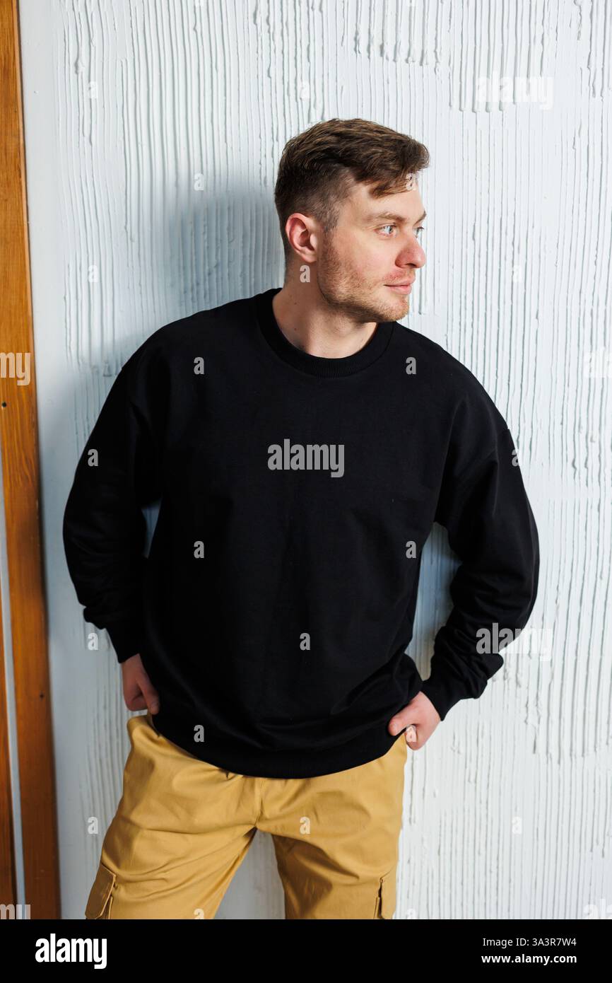 Un uomo che indossa un elegante maglione nero abbinato a pantaloni tan si appoggia in modo informale a una parete con una postura rilassata Foto Stock