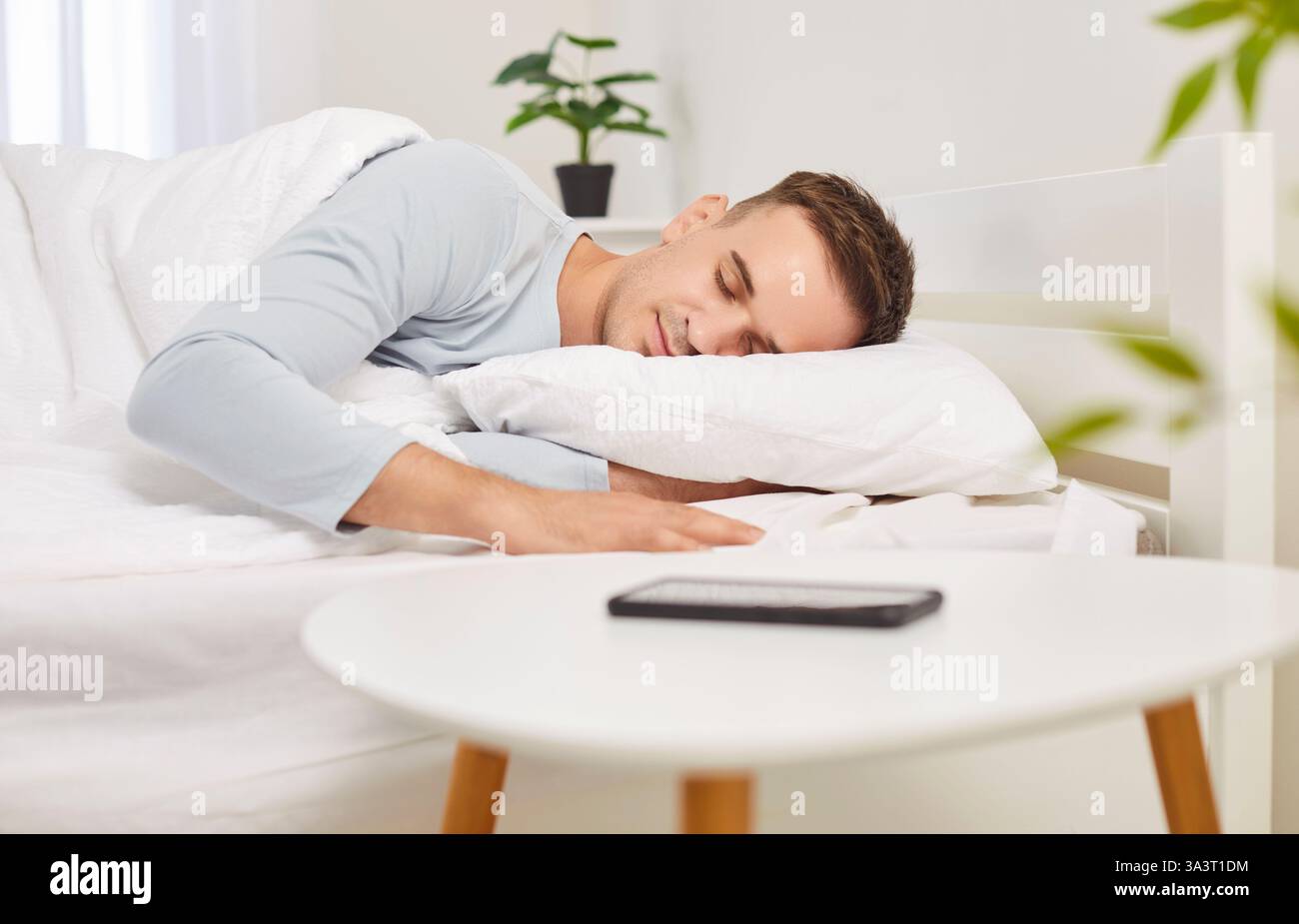 Ritratto di un giovane che dorme sul letto in un'accogliente camera bianca moderna con telefono cellulare sul tavolo. Foto Stock