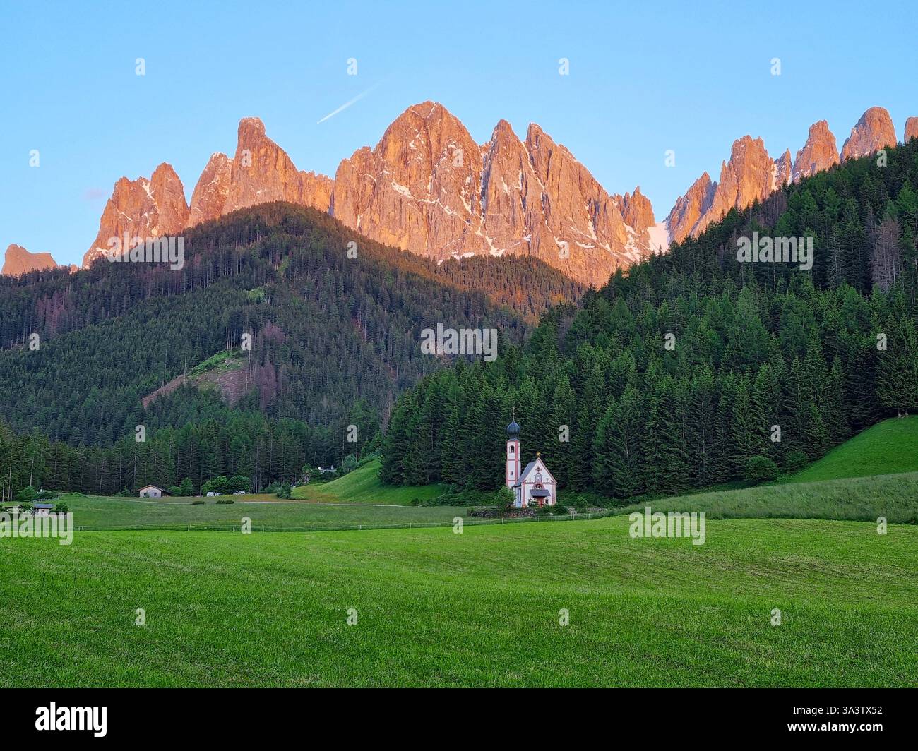 Vista panoramica della famosa cappella di San Giovanni con una cupola a cipolla nel campanile e la cresta di Odle inizia a diventare rossa al tramonto. Dolomiti, Italia Foto Stock