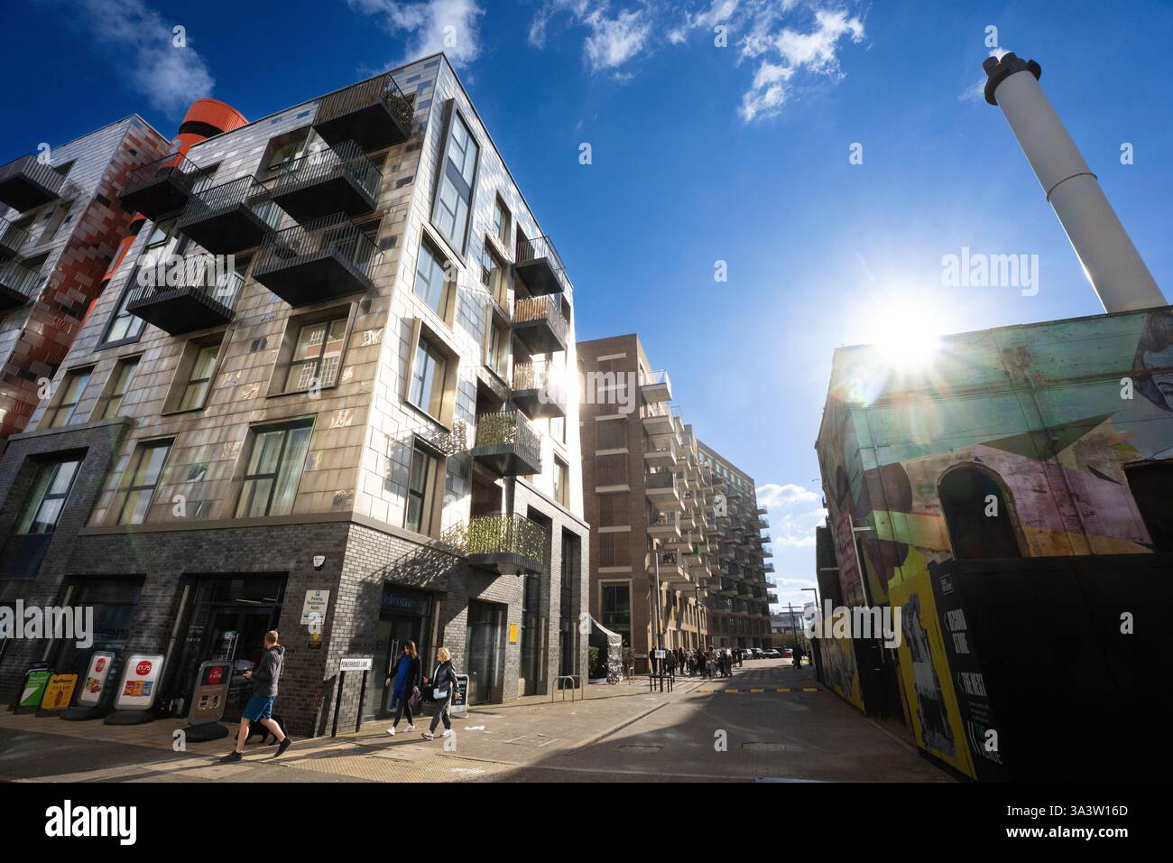 I pedoni camminano lungo Powerhouse Lane, Londra, passando davanti a moderni edifici di appartamenti e a un edificio coperto di murales. Giornata di sole. L'Old Vinyl Factory Festiv Foto Stock