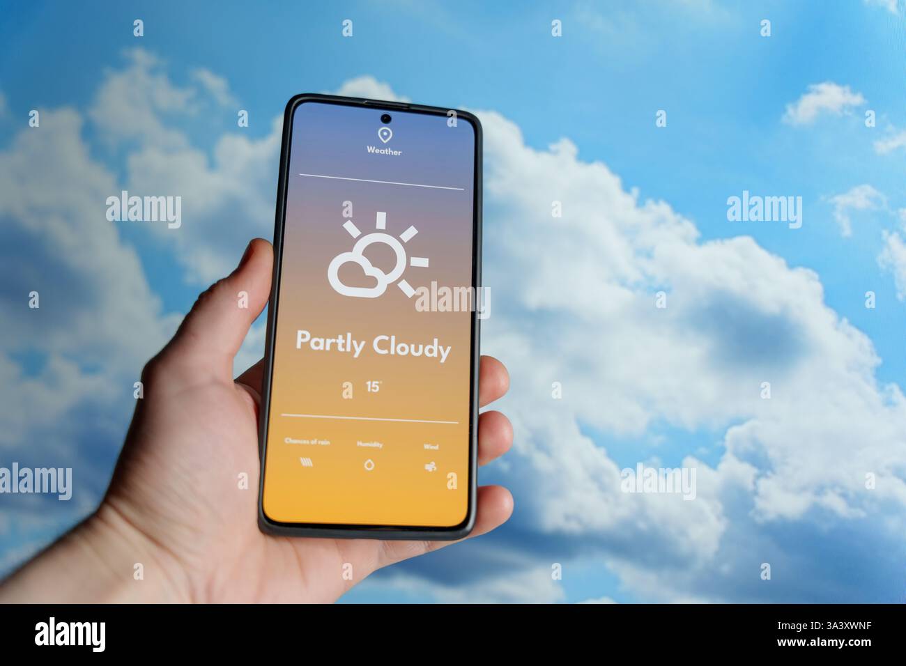 Mano con uno smartphone che visualizza un'app meteo con previsioni parzialmente nuvolose. Cielo blu con nuvole sparse sullo sfondo. Tecnologia meteorologica Foto Stock