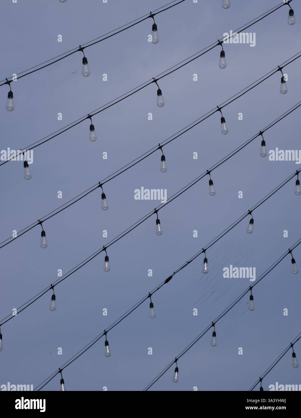 Linee multiple di lampadine appese ai fili in direzione diagonale contro l'alimentazione della tecnologia elettrica del cielo blu e lo sfondo luminoso Foto Stock