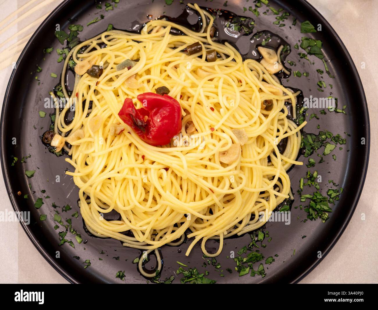Piatto di spaghetti con aglio, olio, peperoncino e prezzemolo servito su un piatto nero guarnito con prezzemolo tritato e pomodoro secco, un classico di i Foto Stock