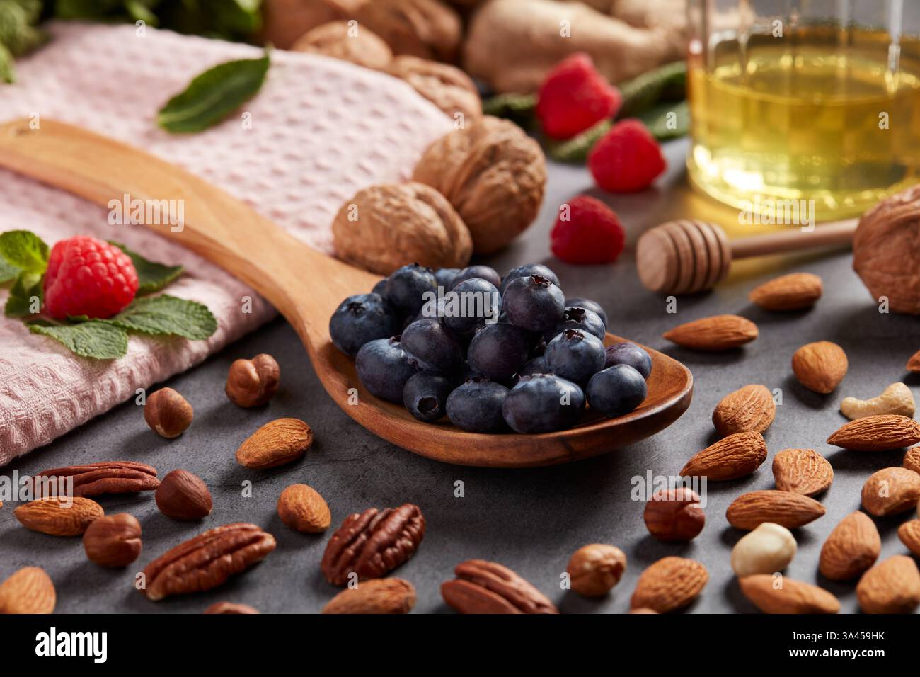 Un cucchiaio di legno pieno di mirtilli si trova su una superficie grigia disseminata di noci, mandorle, nocciole, lamponi e un vaso di miele. Foto Stock