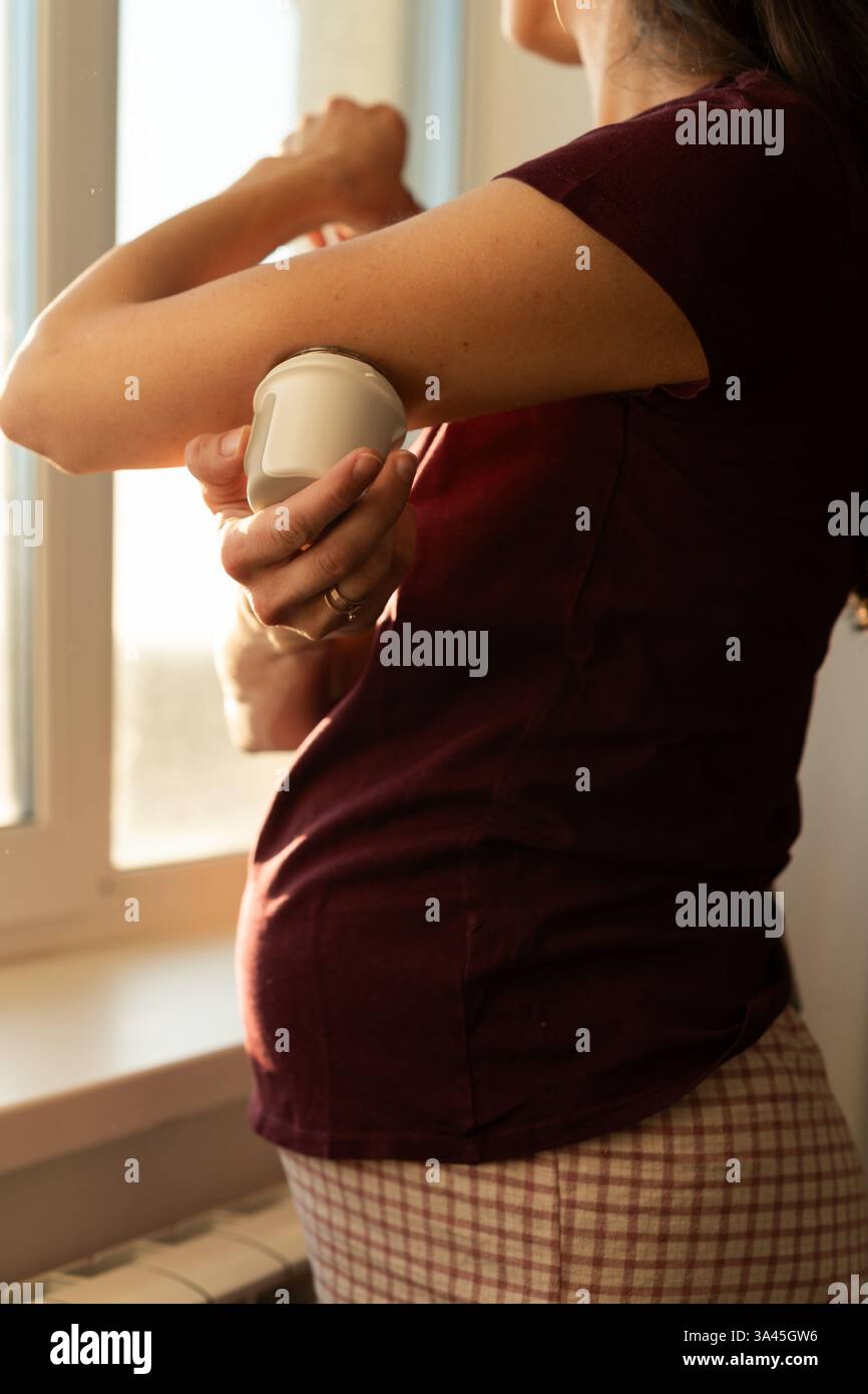 Primo piano di una donna incinta in piedi accanto a una finestra che fissa un monitoraggio continuo del glucosio sul braccio Foto Stock