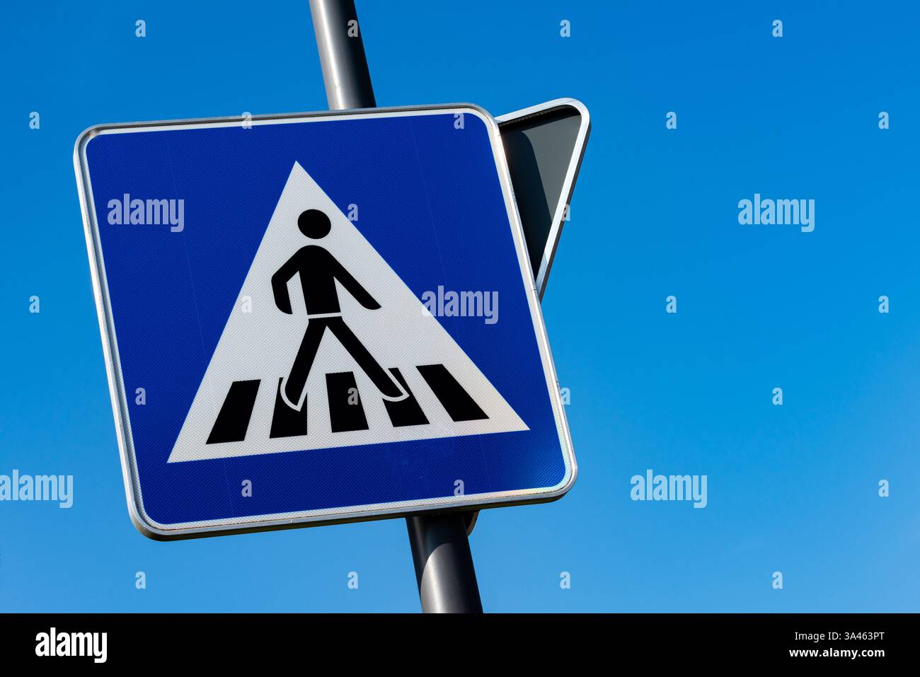 Un cartello triangolare di attraversamento pedonale mostra una figura che cammina su strisce bianche. Il cartello è montato su un palo sotto un cielo azzurro, sottolineando S. Foto Stock