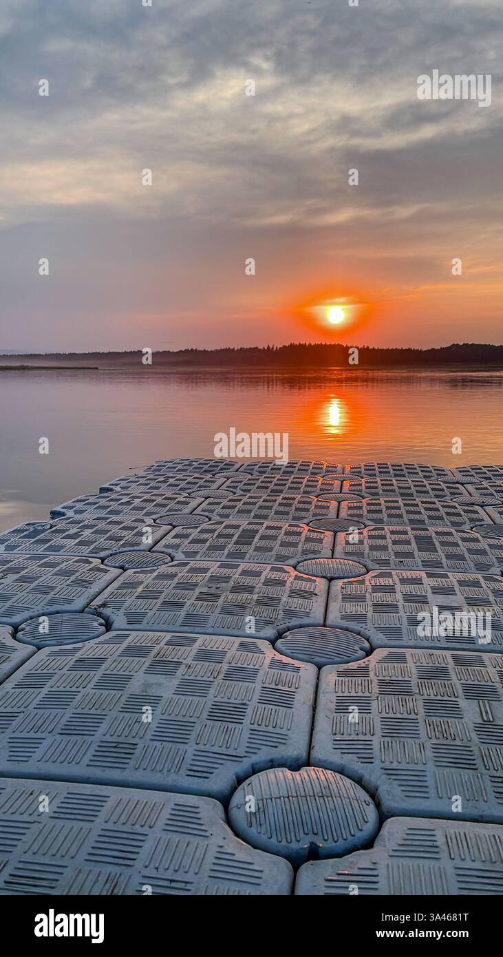 Un molo galleggiante con una superficie strutturata si estende su un lago fermo al tramonto, riflettendo le calde sfumature del cielo sull'acqua. Fotografato in natur Foto Stock