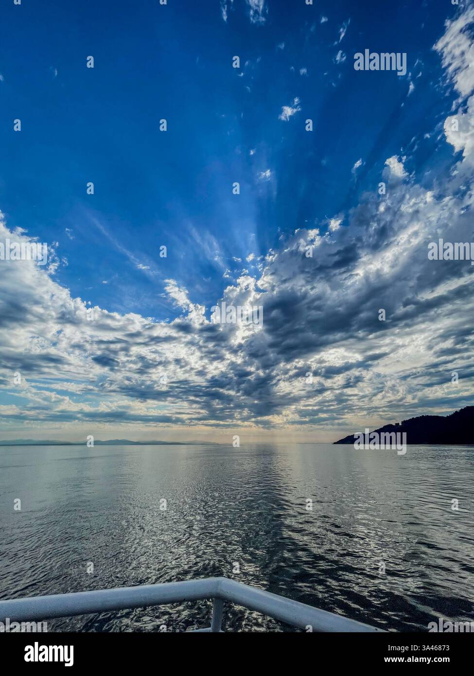 Una vista panoramica dell'oceano catturata da una ringhiera per barche, con acqua testurizzata che riflette il cielo. Nuvole e raggi solari spettacolari aggiungono profondità all'acqua aperta e. Foto Stock