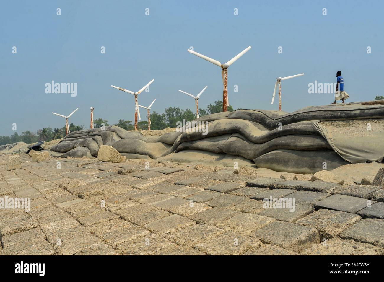 Un uomo locale cammina vicino alle turbine eoliche sulle rive dell'isola di Kutubdia. Un tempo simbolo di speranza per l'energia verde, queste strutture arrugginite ora sono abbandonate. Una volta operativa e generatrice di elettricità, sono cadute in rovina, sollevando preoccupazioni per la cattiva gestione e il futuro delle iniziative del Bangladesh in materia di energie rinnovabili. Isola di Kutubdia, Bangladesh. Foto Stock