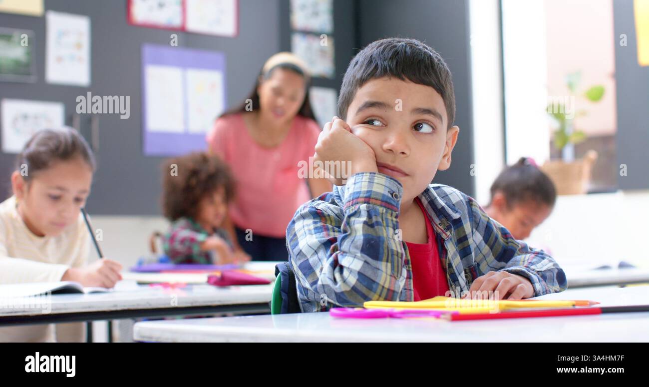 A scuola, ragazzo che sogna ad occhi aperti alla scrivania mentre i compagni di classe e l'insegnante lavorano Foto Stock