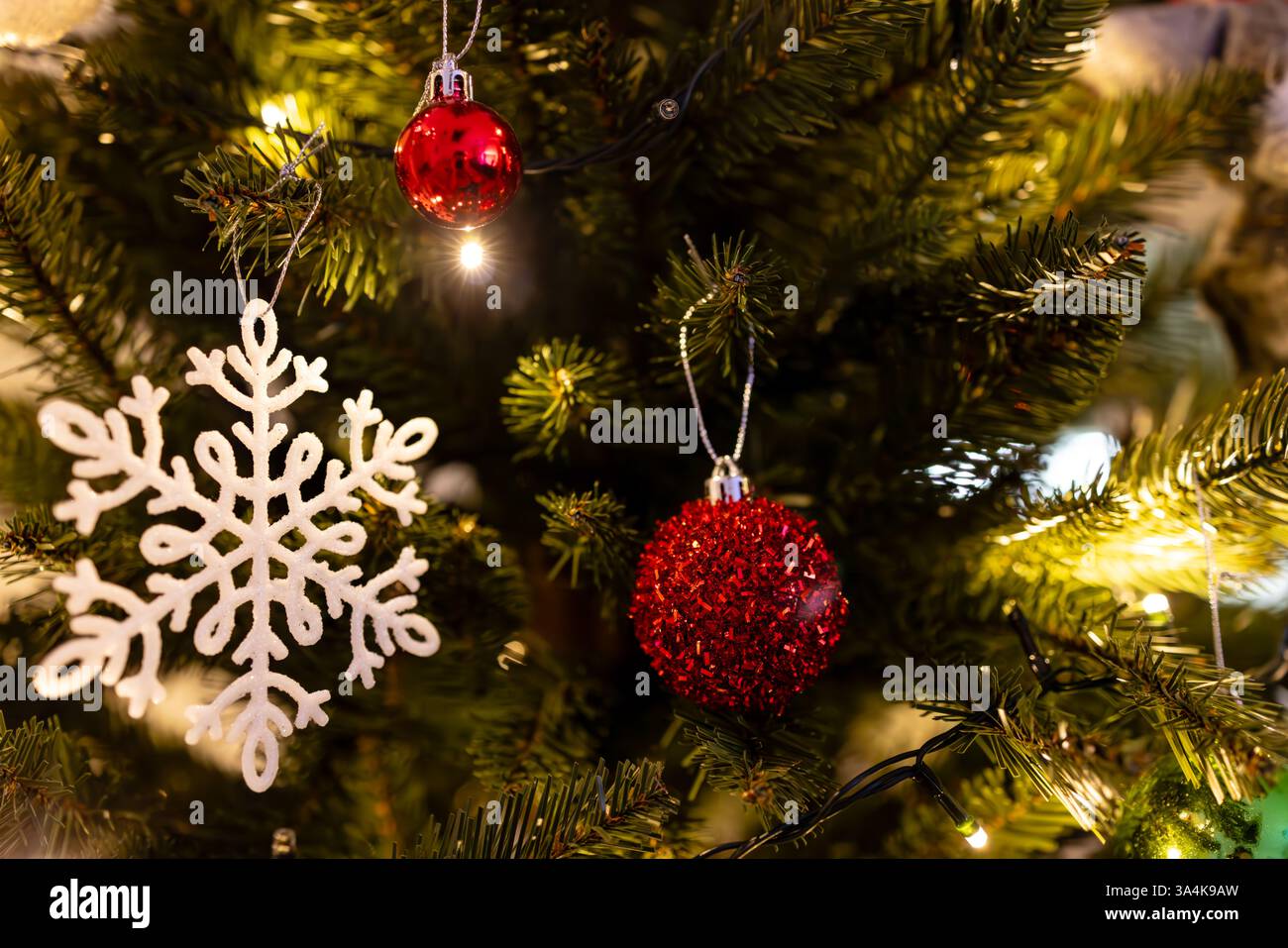 Primo piano di un albero di Natale splendidamente decorato con palline rosse scintillanti, un ornamento bianco con fiocco di neve e luci a corda calde e soffici. Foto Stock