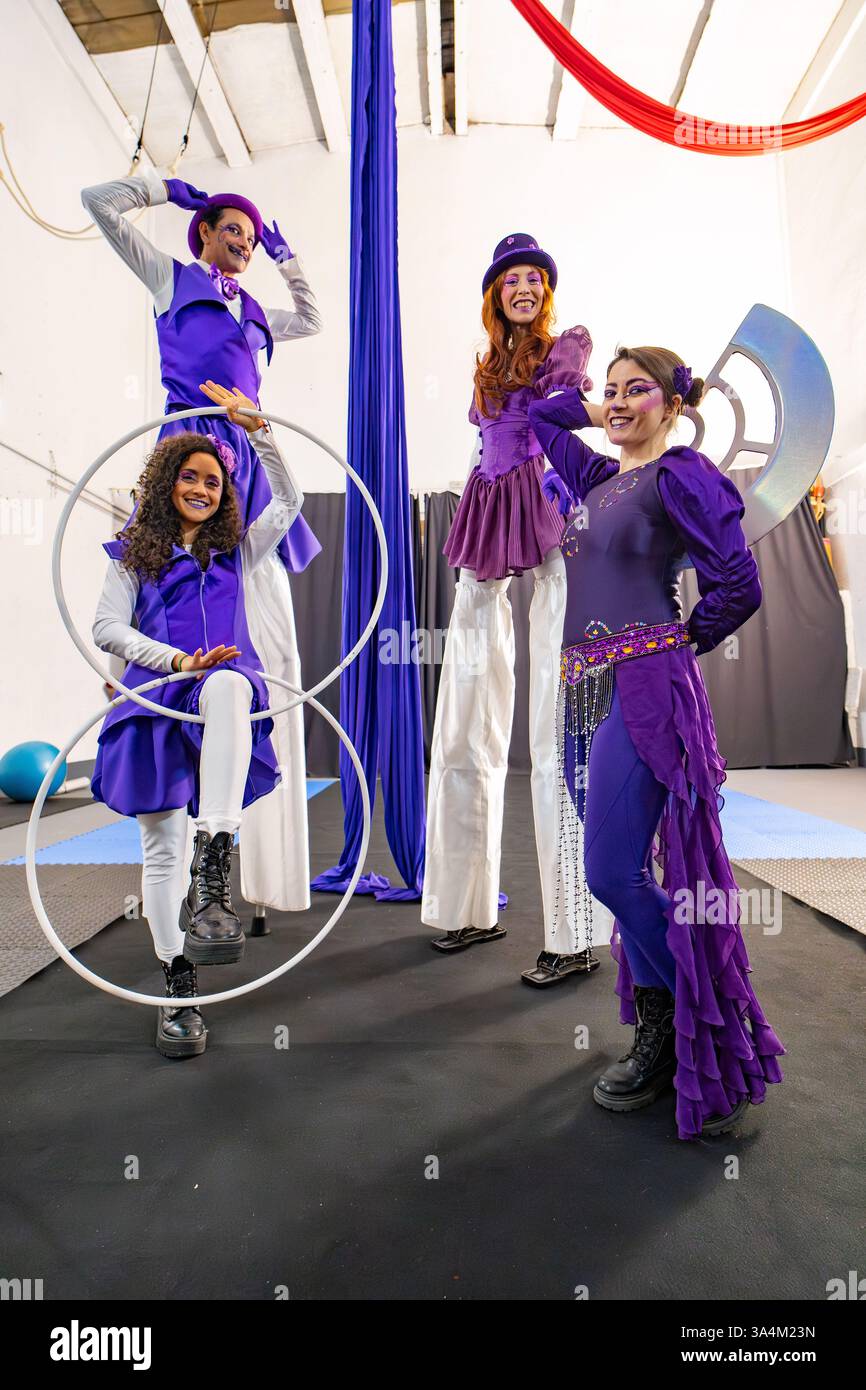 Gli artisti del circo che indossano costumi viola si mettono in posa insieme in un'aula di formazione, mettendo in mostra le loro abilità con trampoli e hula hoops Foto Stock Gli artisti del circo che indossano costumi viola si mettono in posa insieme in un'aula di formazione, mettendo in mostra le loro abilità con trampoli e hula hoops Foto Stock