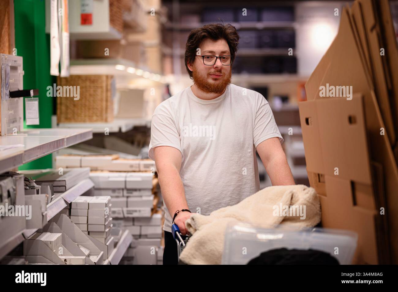 Un uomo con gli occhiali e la barba spinge un carrello pieno di oggetti essenziali per la casa in un corridoio di un negozio, circondato da contenitori e oggetti per la casa. Foto Stock