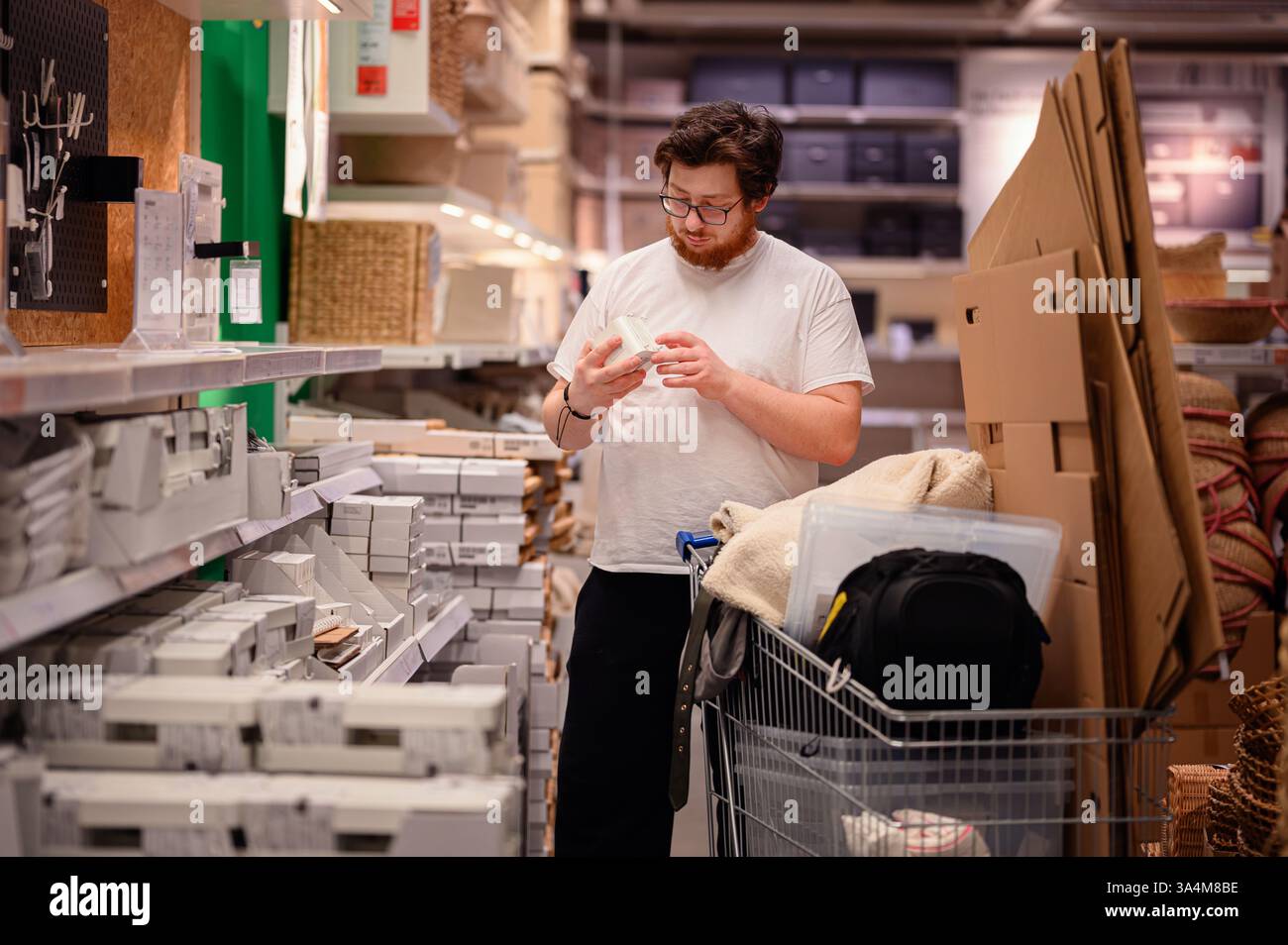 Un uomo con una t-shirt bianca esamina un prodotto mentre acquista gli articoli essenziali per la casa. Si trova vicino a scaffali pieni di oggetti per la casa, con un carrello pieno Foto Stock