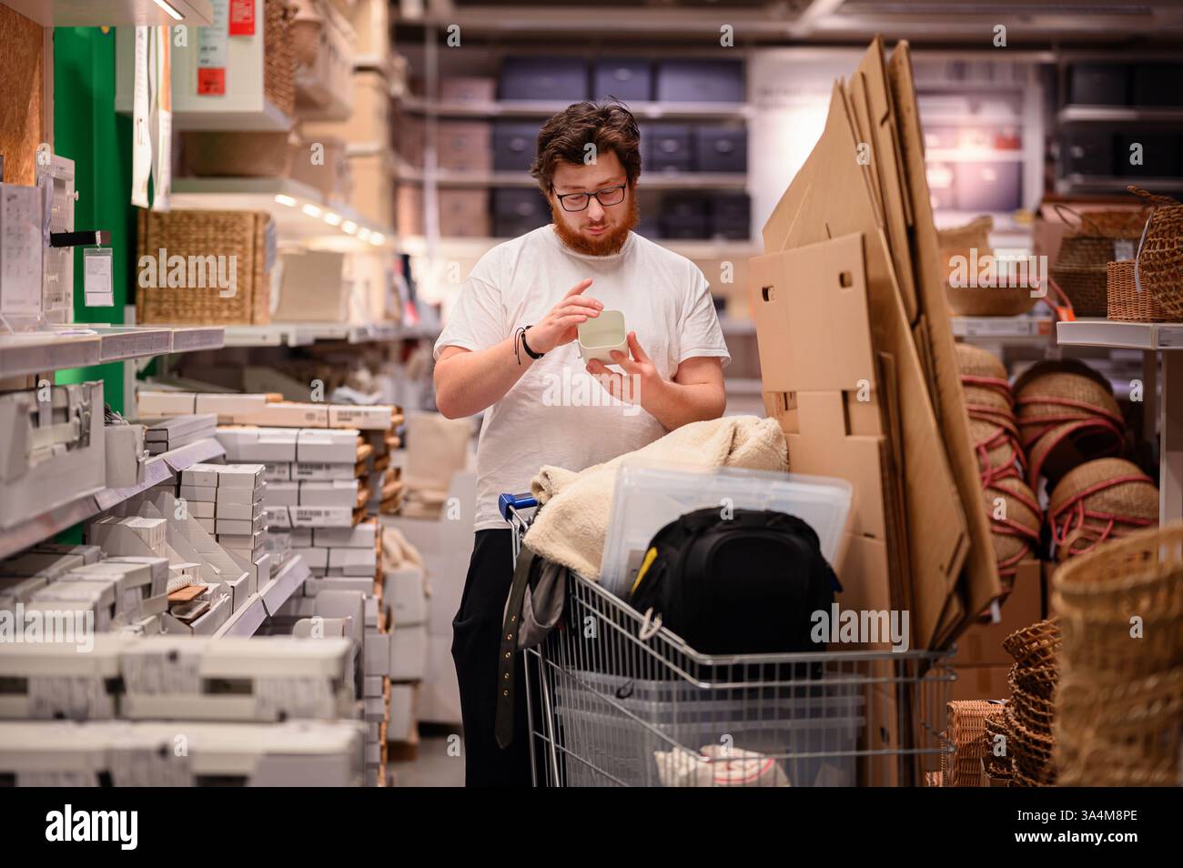 Un uomo con una t-shirt bianca esamina un prodotto mentre acquista gli articoli essenziali per la casa. Si trova vicino a scaffali pieni di oggetti per la casa, con un carrello pieno Foto Stock