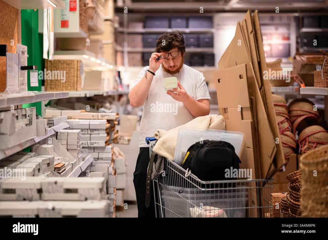 Un uomo con una t-shirt bianca esamina un prodotto mentre acquista gli articoli essenziali per la casa. Si trova vicino a scaffali pieni di oggetti per la casa, con un carrello pieno Foto Stock