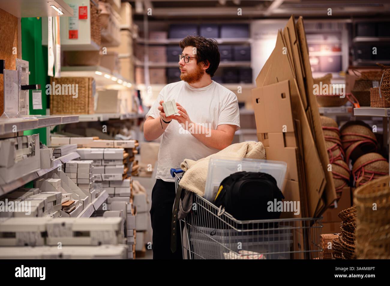 Un uomo con una t-shirt bianca esamina un prodotto mentre acquista gli articoli essenziali per la casa. Si trova vicino a scaffali pieni di oggetti per la casa, con un carrello pieno Foto Stock