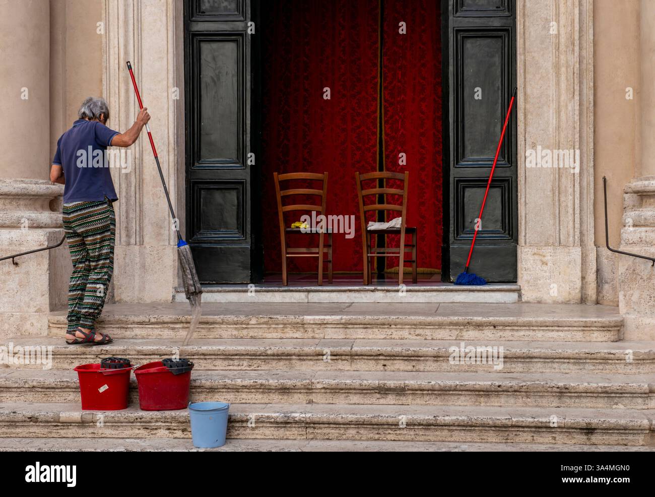 Una signora che indossa pantaloni colorati pulisce i gradini di una vecchia chiesa nel centro di Roma con secchi e scope rosse, mentre due sedie vuote vengono utilizzate per contenere le attrezzature per la pulizia. Foto Stock