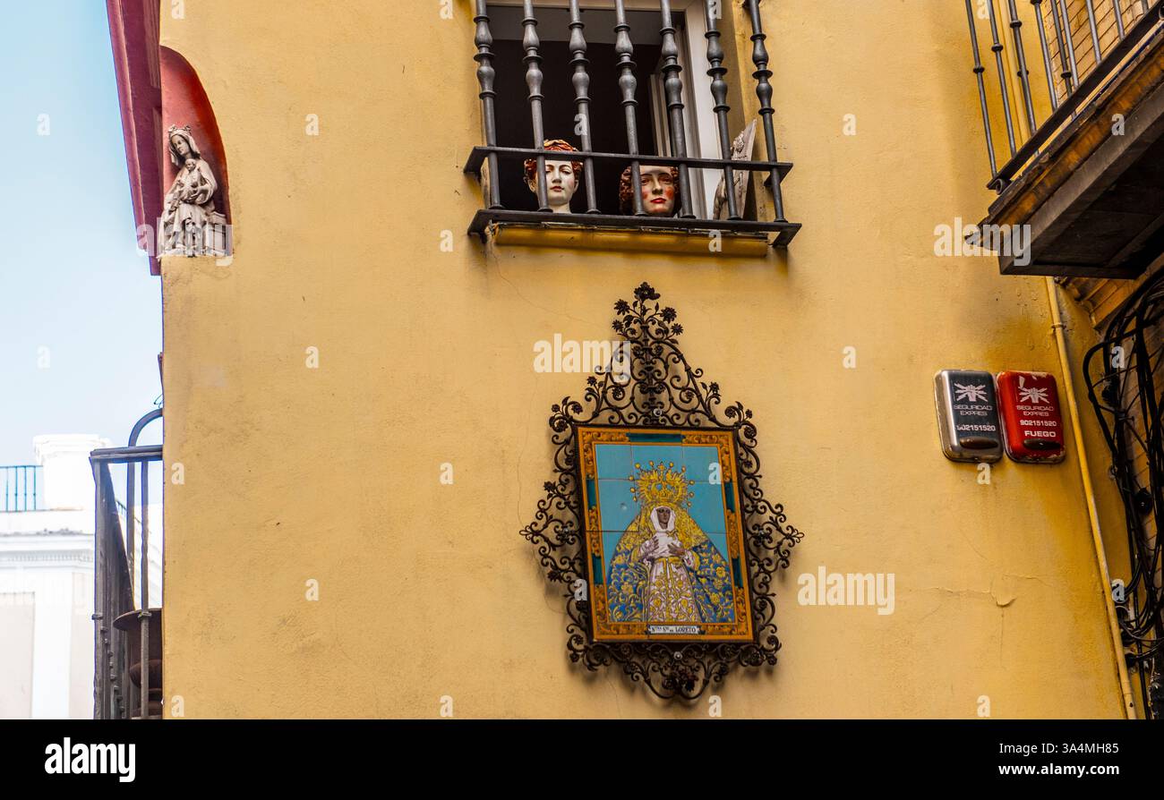 Le colorate teste di figure sbirciano fuori da un balcone adornato da un'impressionante opera religiosa su una parete gialla illuminata dal sole che mostra la cultura locale e l'espressione artistica a Siviglia in Spagna. Foto Stock