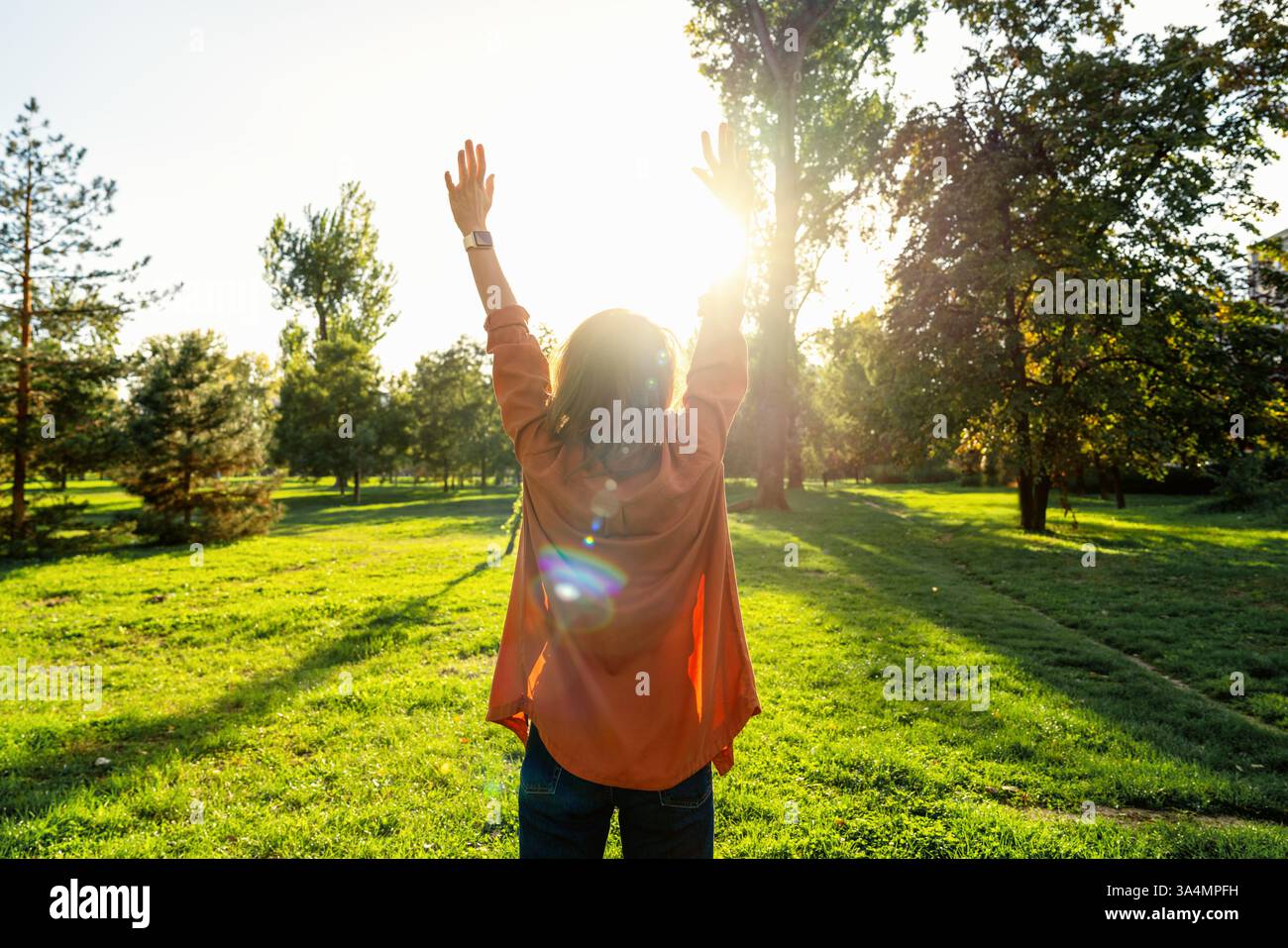 Vista posteriore della donna con le braccia alzate in piedi davanti al verde, sentendosi libera e felice mentre si gode la natura. Foto Stock