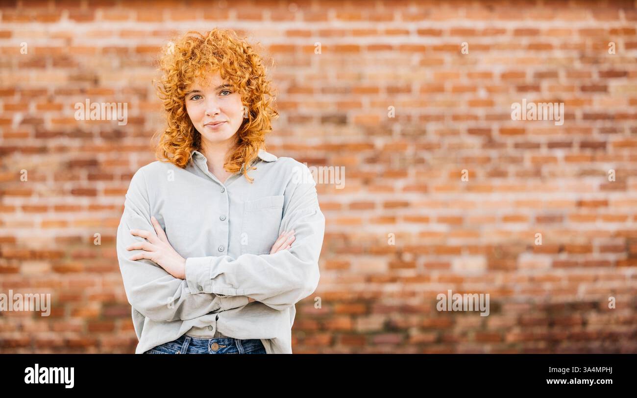 Ritratto di una giovane donna rossa sicura di sé con le braccia incrociate in posa davanti a un muro di mattoni Foto Stock