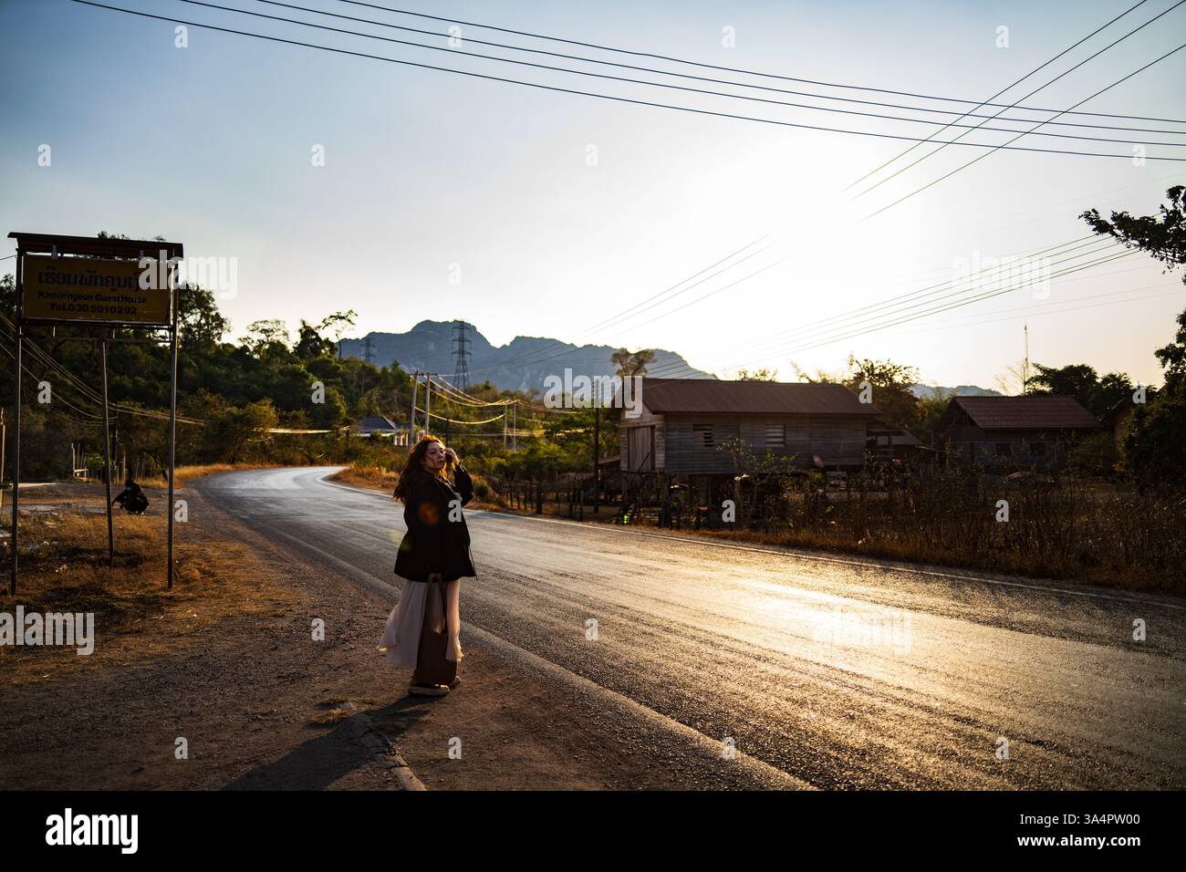 Vista panoramica di una strada rurale al tramonto nel sud-est asiatico con un viaggiatore che scatta foto, mostrando case tradizionali in legno e vegetazione lussureggiante Foto Stock