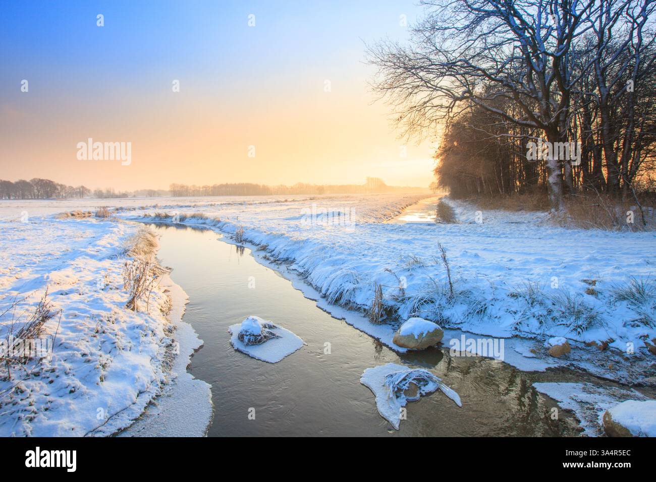 Un vibrante paesaggio invernale con un campo coperto di neve e un fiume che scorre attraverso il paesaggio con un cielo colorato e una luce solare dorata Foto Stock Un vibrante paesaggio invernale con un campo coperto di neve e un fiume che scorre attraverso il paesaggio con un cielo colorato e una luce solare dorata Foto Stock