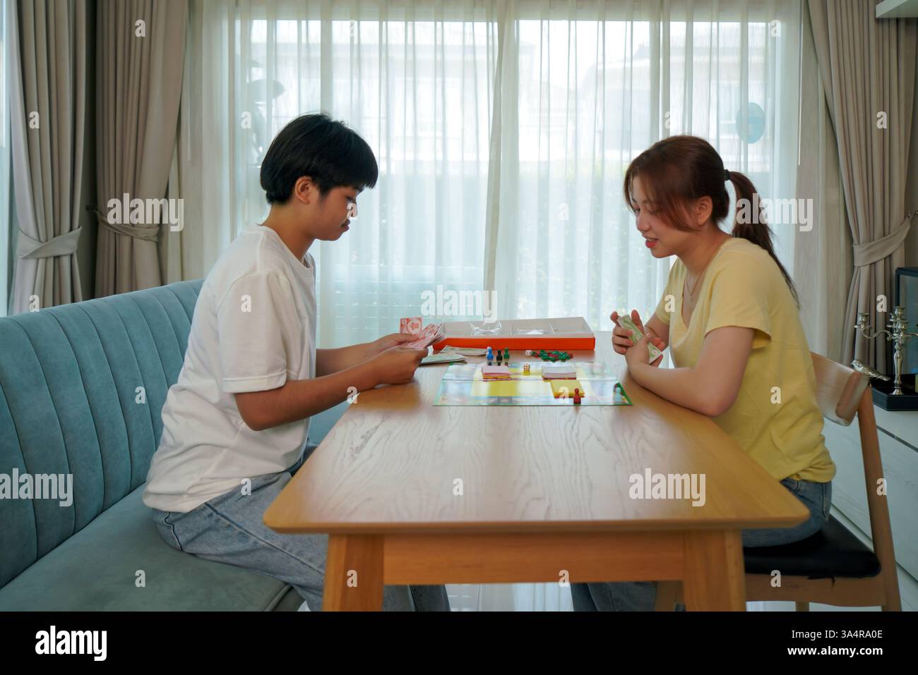 Due amici asiatici giocano il classico gioco da tavolo Monopoli ad un tavolo di legno. Uno sta smistando attraverso il denaro monopolistico, mentre l'altro sta pianificando la strategia Foto Stock