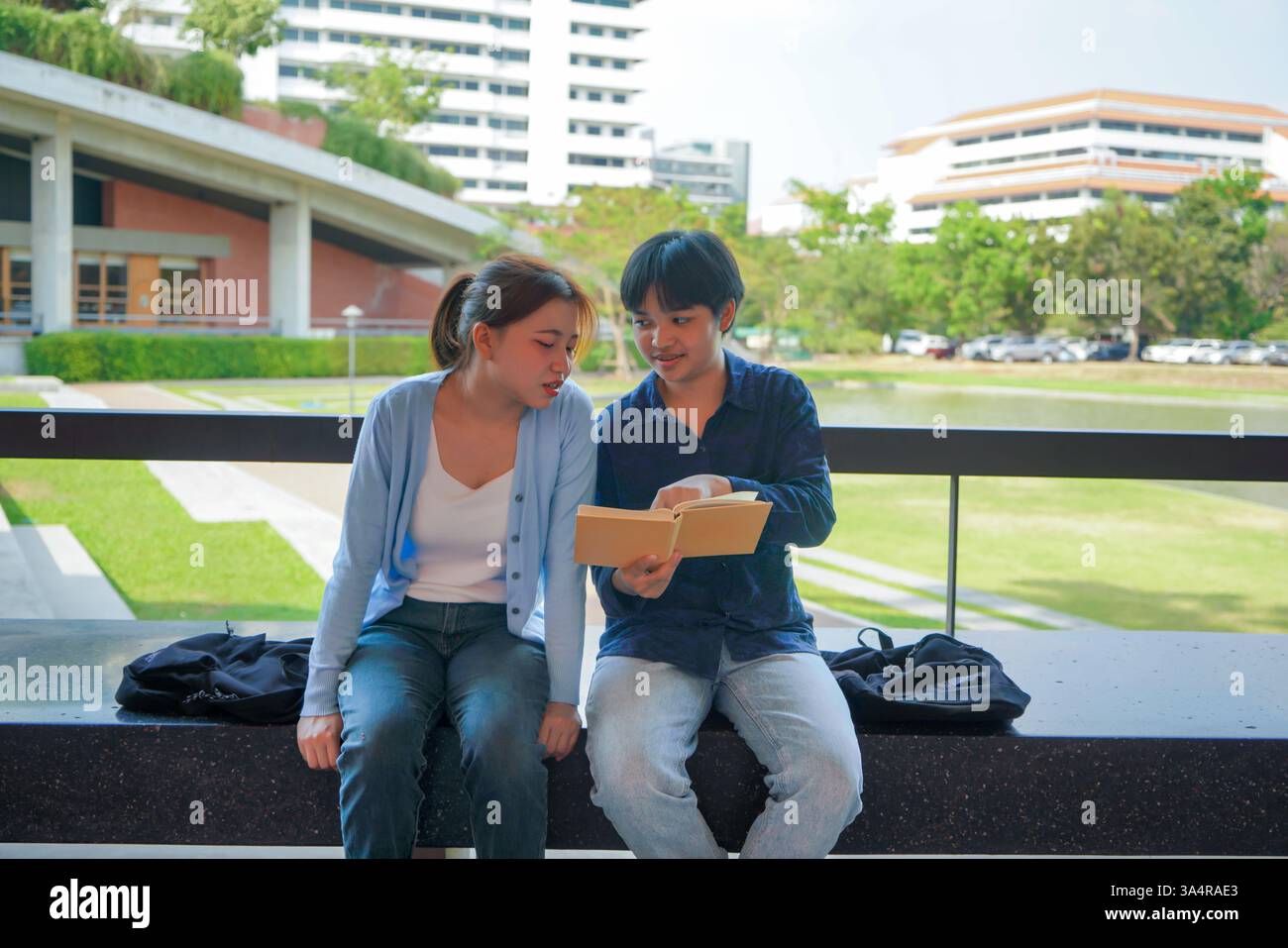 due studenti universitari asiatici seduti su una panchina in un ambiente universitario moderno, impegnati in una discussione amichevole su un libro condiviso Foto Stock