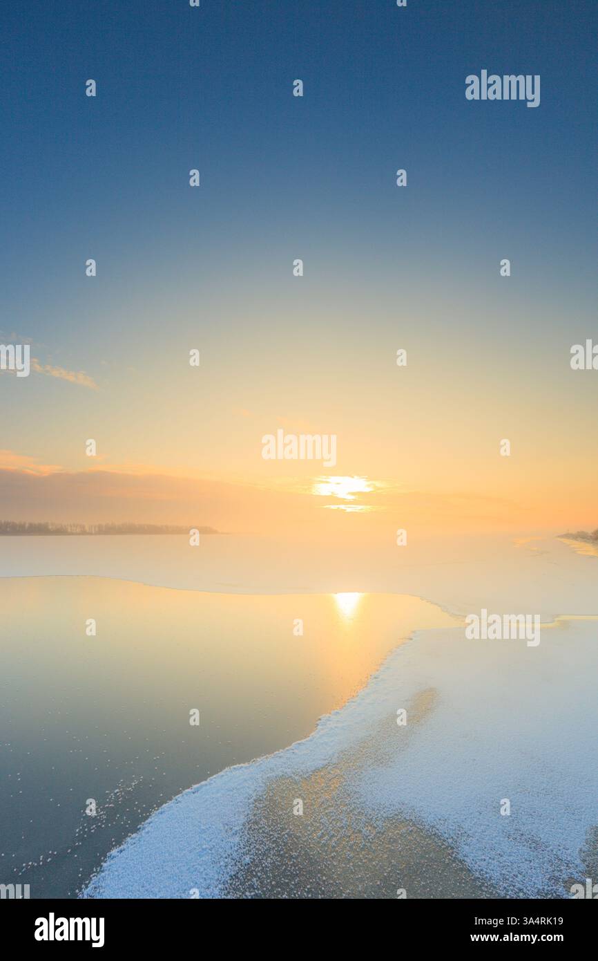 Un paesaggio ghiacciato con un lago coperto di neve all'alba con una calda luce solare nel cielo in una fredda giornata invernale a dicembre Foto Stock Un paesaggio ghiacciato con un lago coperto di neve all'alba con una calda luce solare nel cielo in una fredda giornata invernale a dicembre Foto Stock
